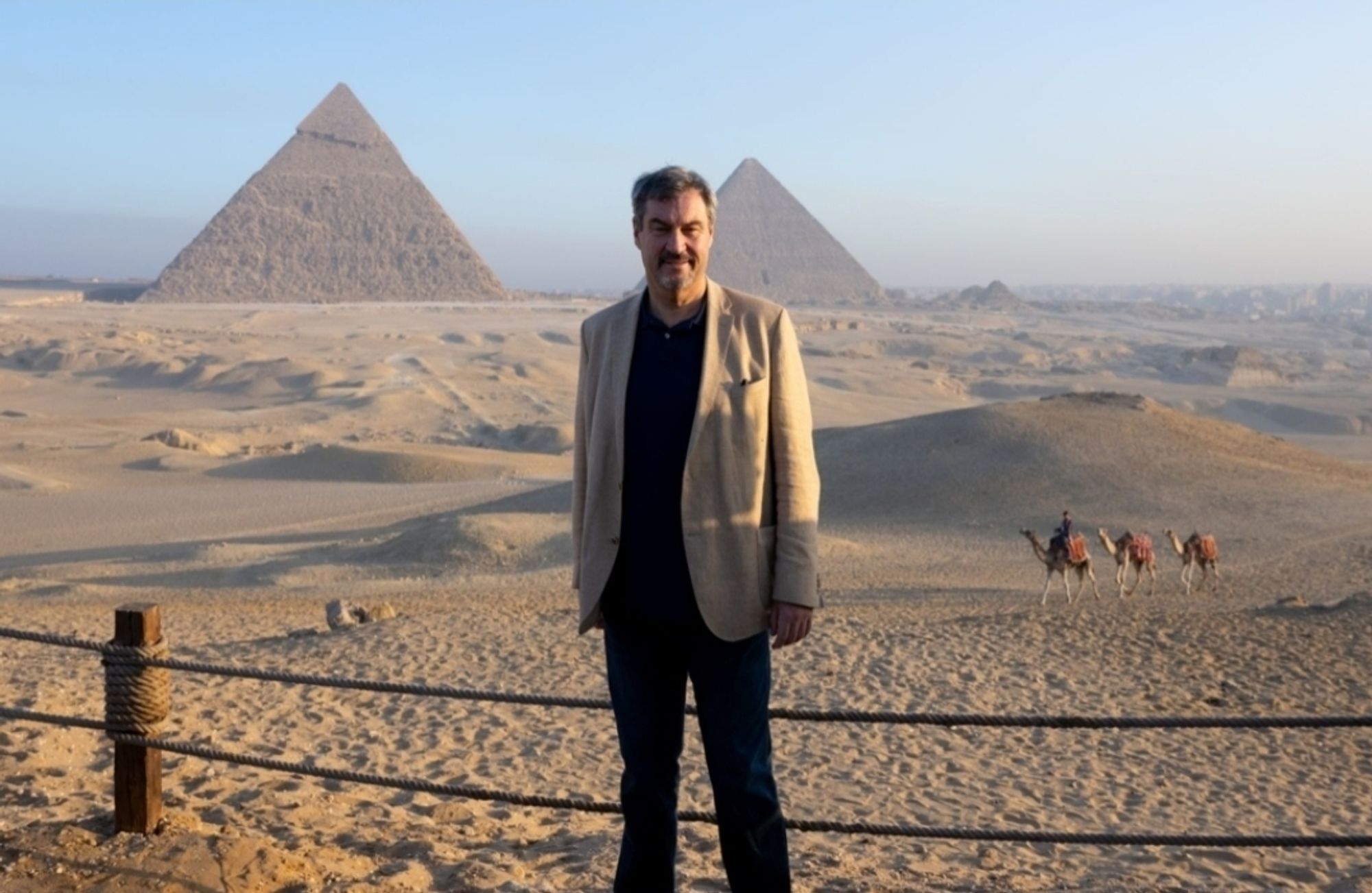 Markus Söder in der Wüste vor den Pyramiden von Gizeh. Eine Person reitet auf einem Kamel mit 2 weiteren Kamelen im Schlepotau durchs Bild.