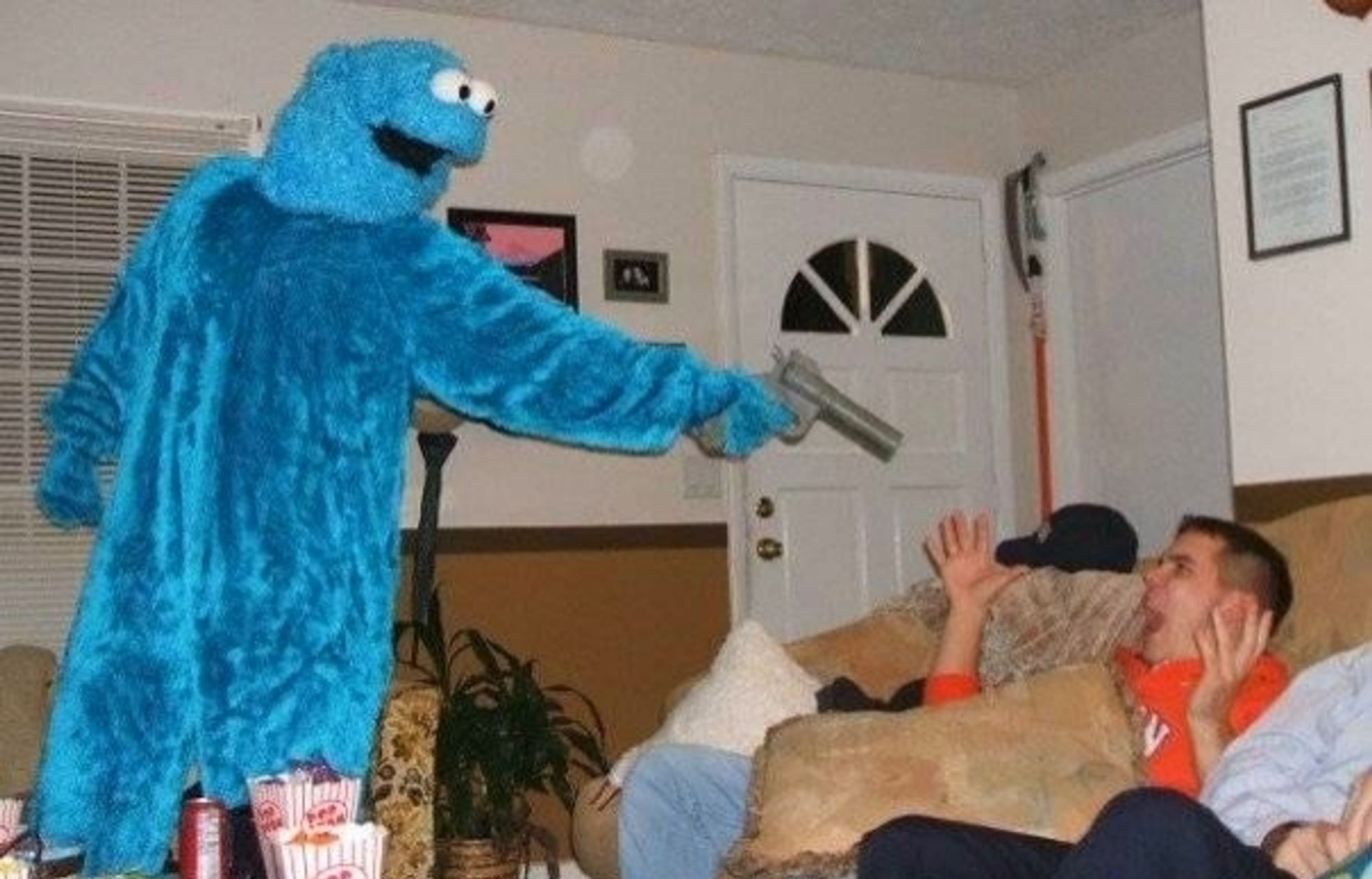 Jemand in einem Cookie Monster Kostüm steht mit einer Spielzeugpistole vor einer Couch auf der ein Mann verängstigt schreiend die Hände in die Luft hält