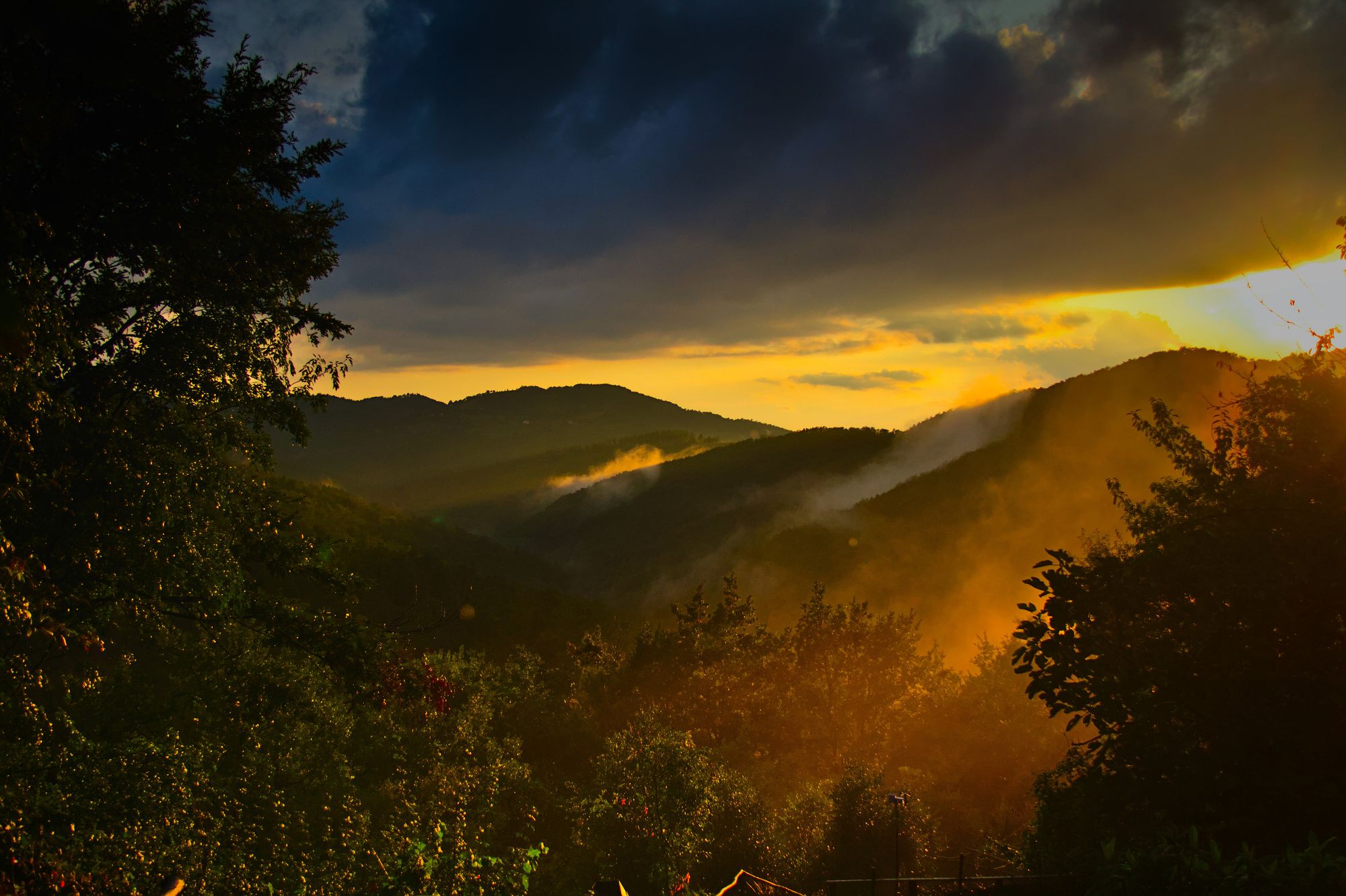 a dark misty valley illuminated in orange light by the setting sun on the right of the frame under a dark cloudy sky