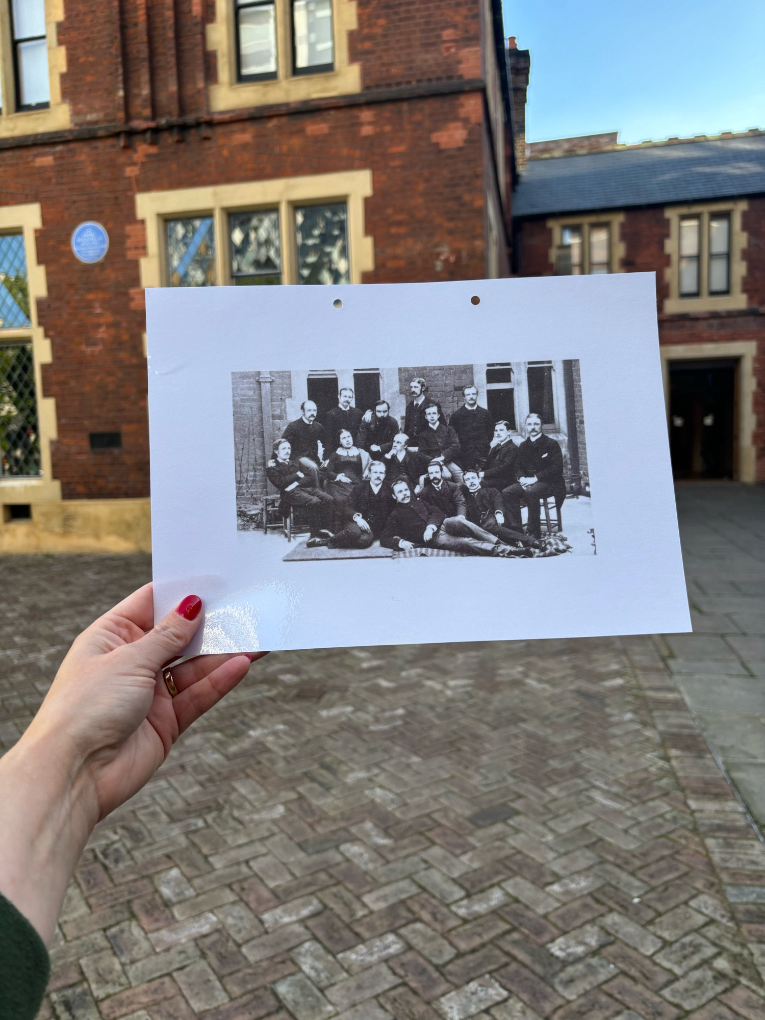 White woman holding up a picture of Victorian men and a woman. Behind is a Victorian building