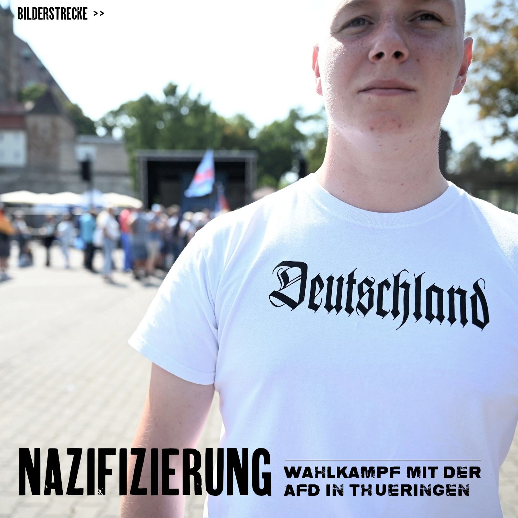 Ein junger Mann mit weißem Shirt steht im Vordergrund. Im Hintergrund sind Menschen vor einer Bühne zu sehen. Die hintere Szenerie ist unscharf. Auf dem Shirt des Mannes ist in Frakturschrift der Aufdruck Deutschland in schwarzer Farbe zu sehen.