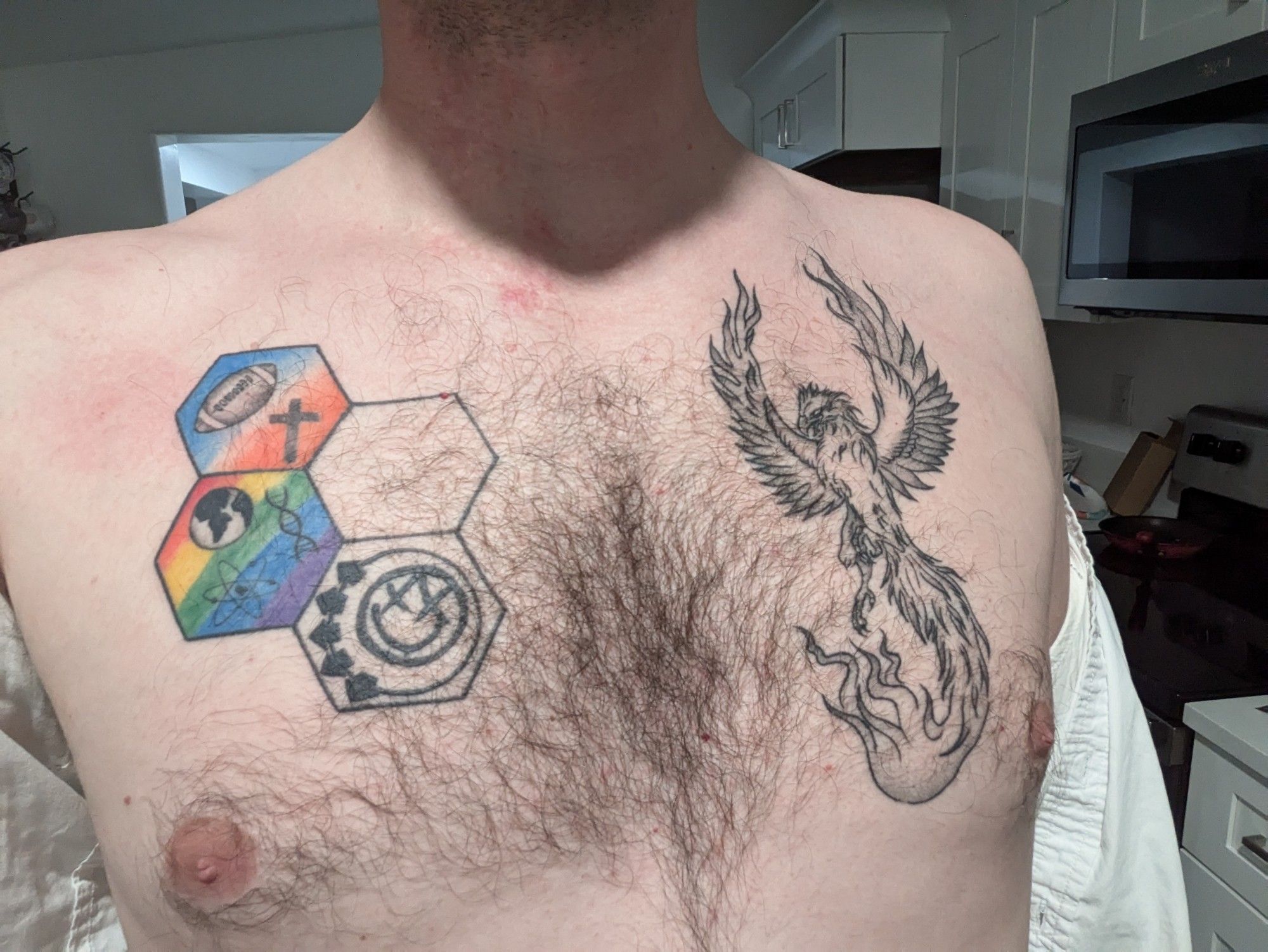 Bare chest of a man with a button up shirt hanging off his arms on the side. On his left is a tattoo of a phoenix, on his right is a tattoo of four connected hexagons. One has a blue and orange background with a football and a cross, one is empty, one has a rainbow background with a silhouette of a globe, a DNA double helix, and an atomic model, and the final with no background and the Blink 182 smiley face symbol.