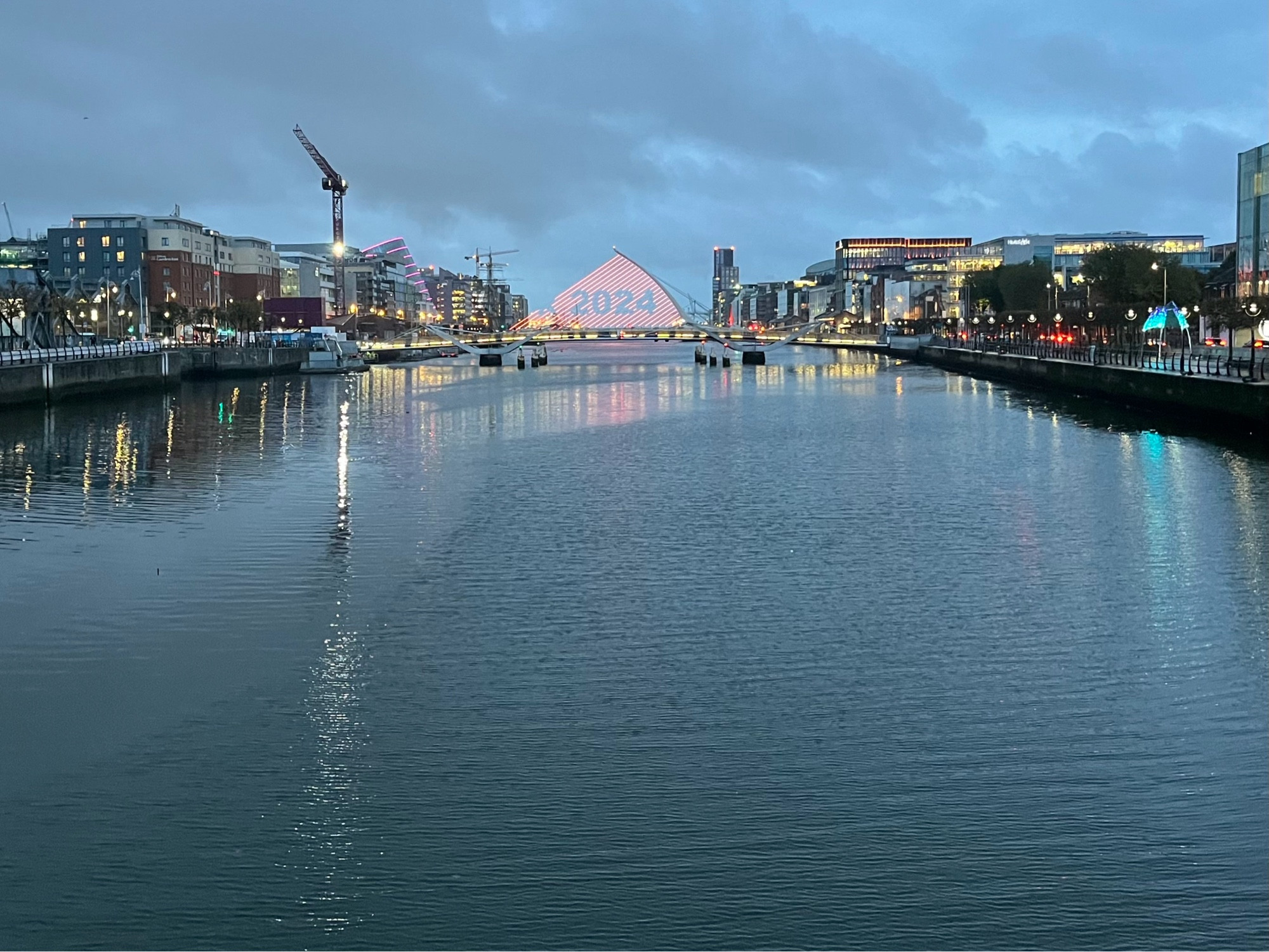 View of the Liffey from Talbot Bridge: the lights from buildings at either side, and even a tall crane whose reflection stretches to the bottom of the picture frame, do not meet in the middle, and the dark power of this broad, deep tidal river can be judged by the ripples of current in the middle. Central and in the top-third of the frame, the Sam Beckett Bridge, still lit in the grey dawn light, shines red along the stands of its harp-shaped back, creating a canvas with the numbers 2024 picked out from the colour.