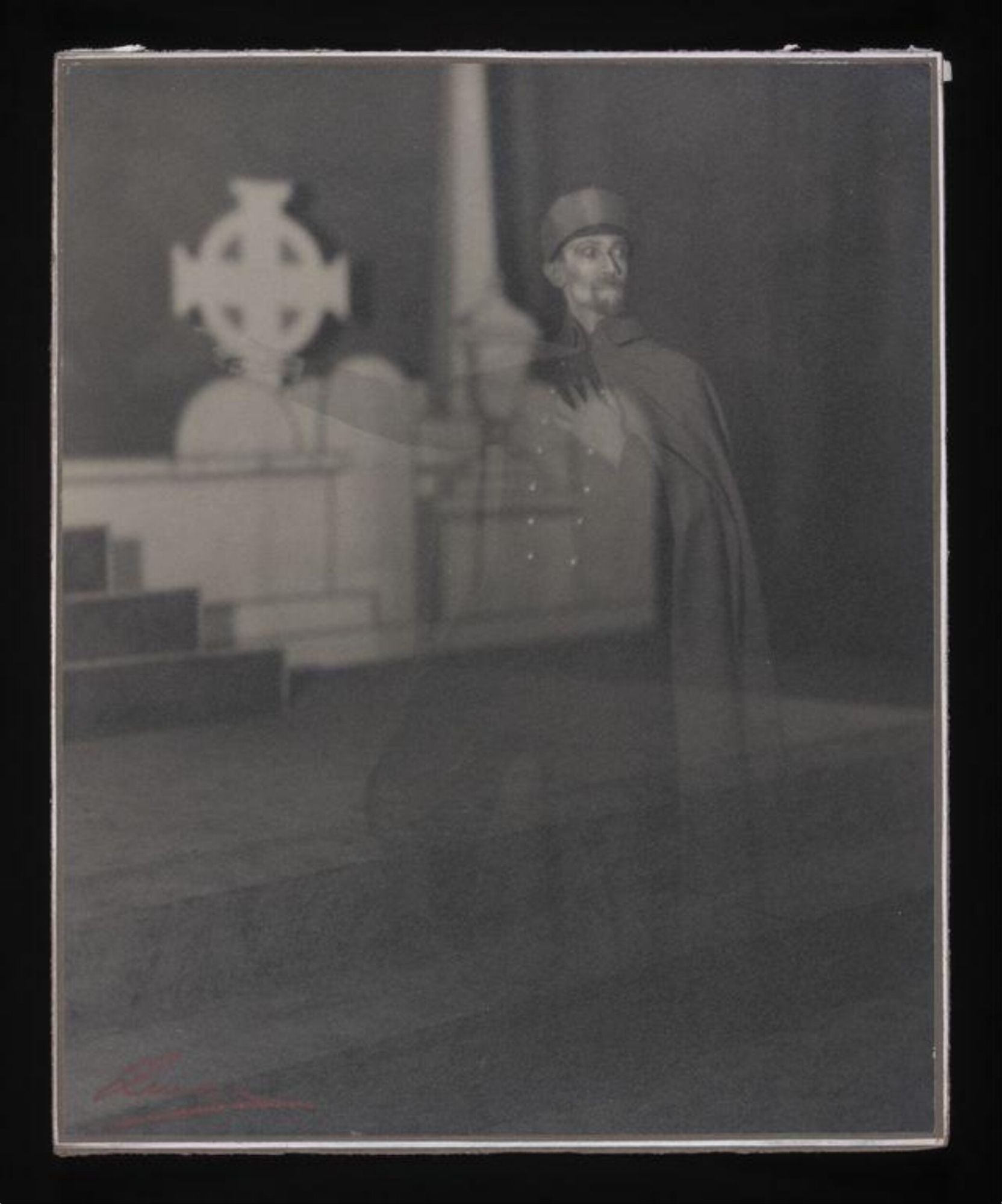 A black and white photograph of a play in performance. In the background there are white tombstones, including a Celtic cross. In the foreground, but transparent, is a man looking to his left, his left hand on his heart, his right hand pointing out of the frame. He is wearing army uniform with a long cloak and cap and has a short moustache and beard.