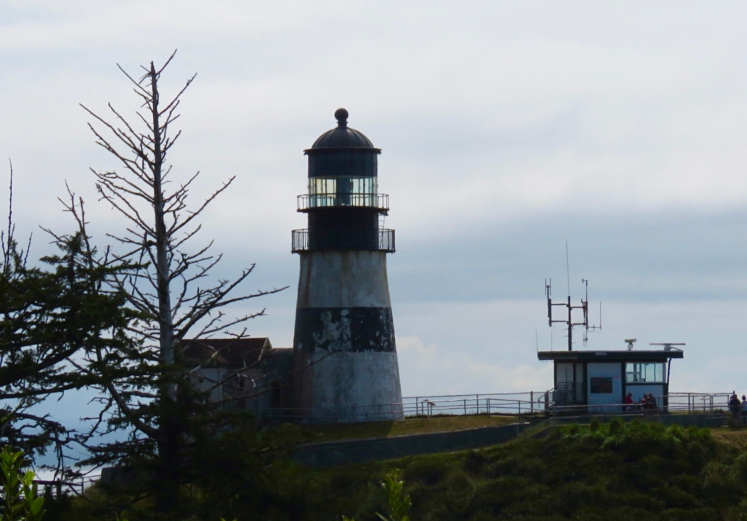 the Cape Disappointment Lighthouse is the oldest operating lighthouse in the PNW. it is in dire need of restoration. we visited on a very dreary day in 2022. there is a building with antennas on the roof to the right and bare trees on the left. 