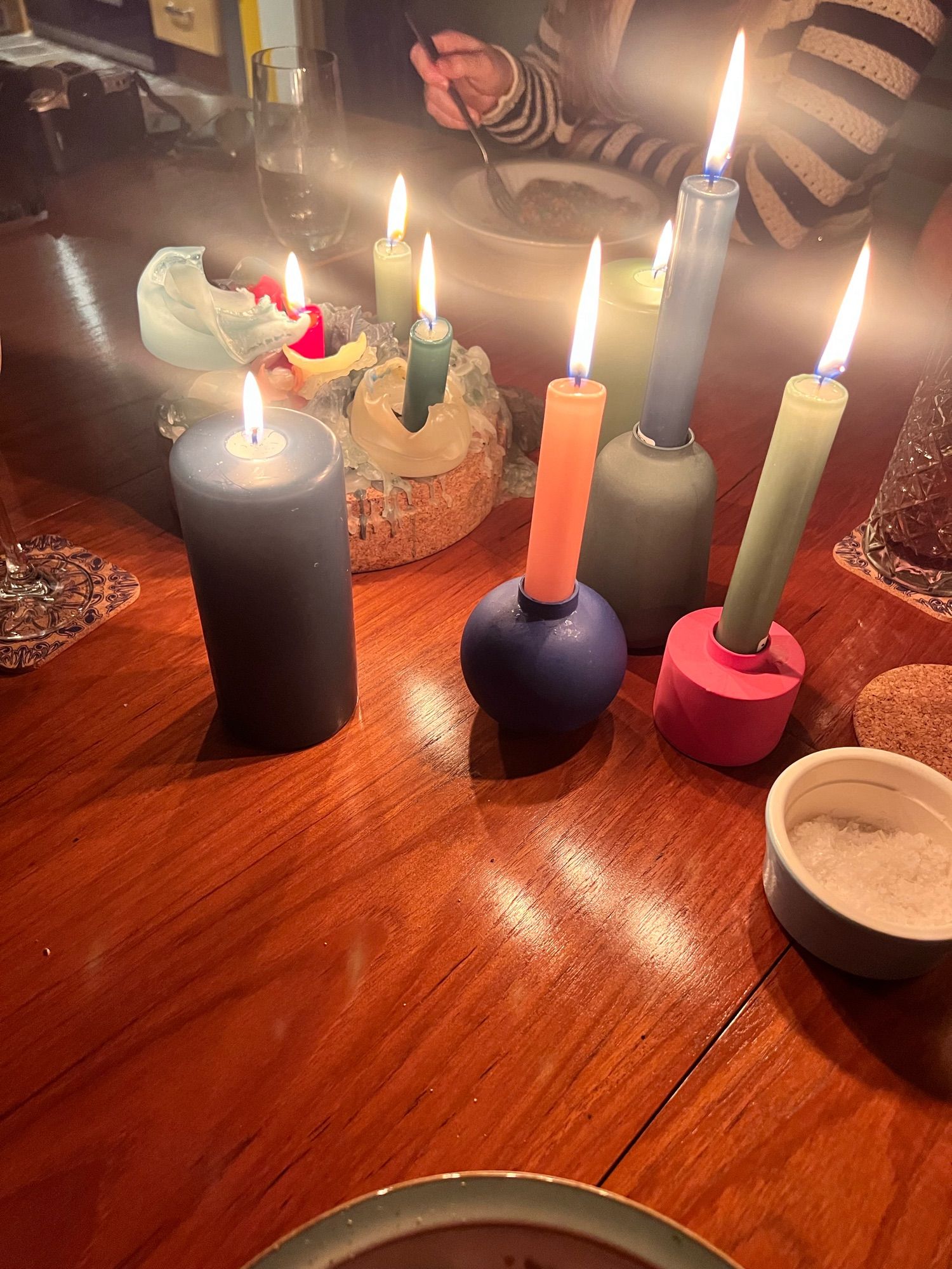 Multiple candles of different colour on a wooden table. The wood is rich and dark. A white and black jumper is in view of someone sitting at the table.