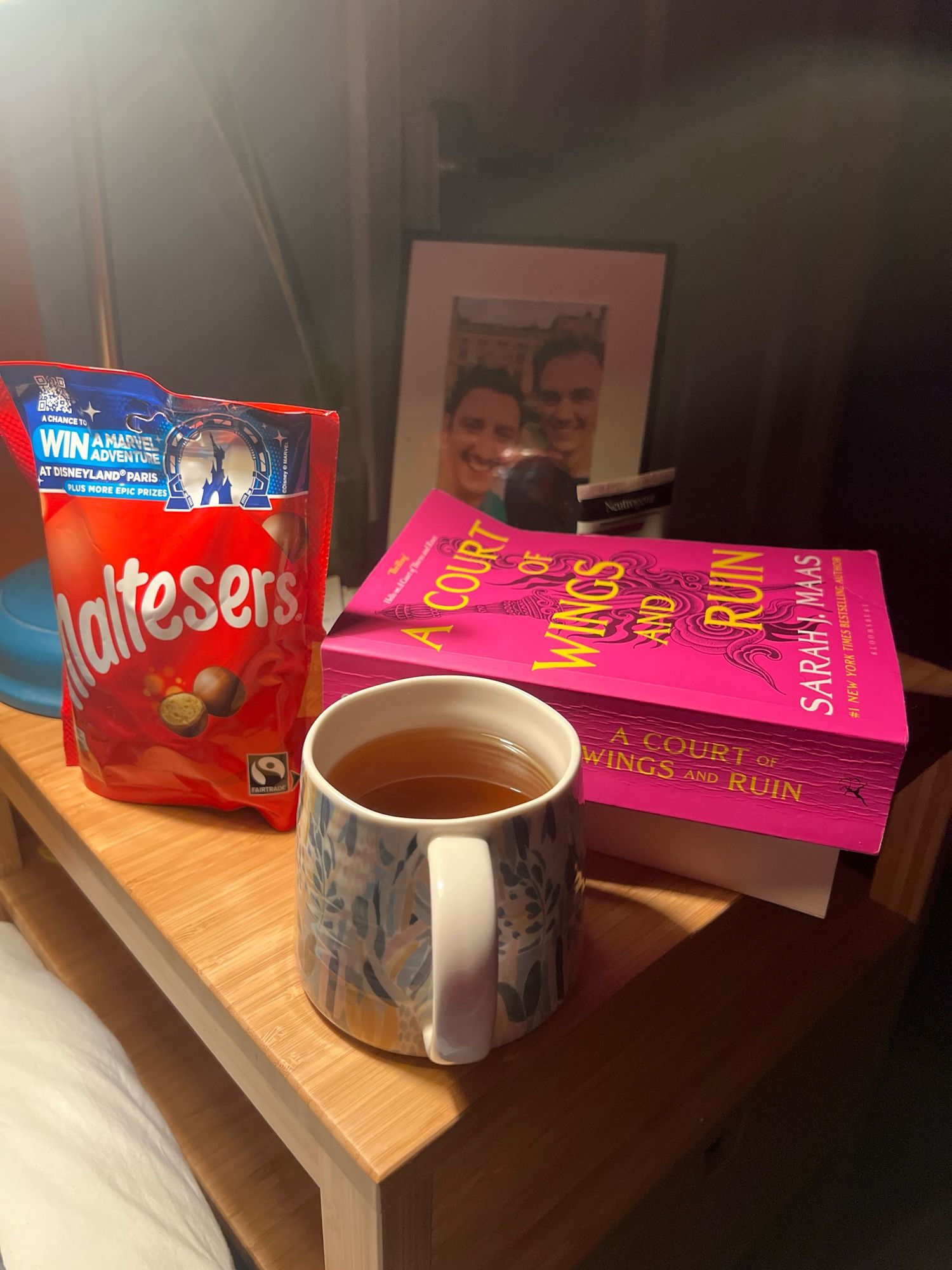 A mug of mint tea, a pink book and a bag of malteasers on a bedside table lit by a lamp