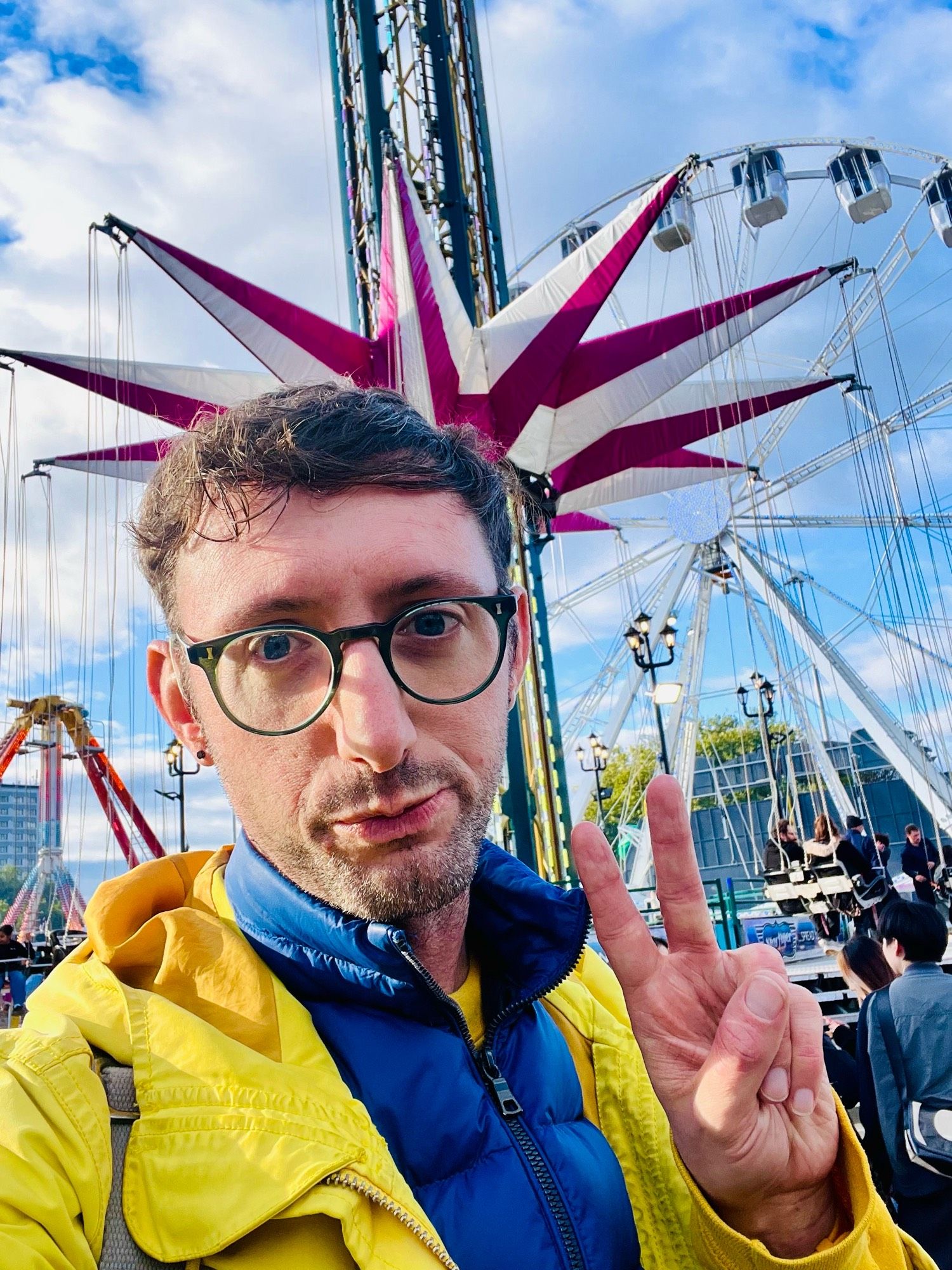 Jake in a yellow and blue jacket makes a v sign. Behind fairground rides.