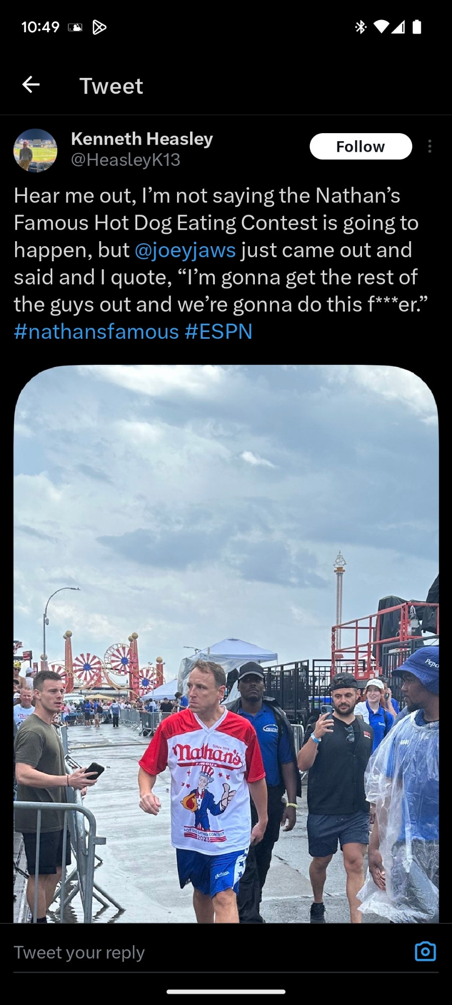 Screenshot of a tweet from user HeasleyK13 saying "Hear me out, I'm not saying the Nathan's Famous Hot Dog Eating Contest is going to happen, but @joeyjaws just came out and said and I quote, "I'm going to get the rest of the guys out and we're going to do this f***er". #nathansfamous #espn". The tweet is accompanied by a photo of Joey Chestnut, who appears to be thoroughly drenched by rain, accompanied by an entourage of various people walking along the sidewalk with the Coney Island amusement park in the background. 