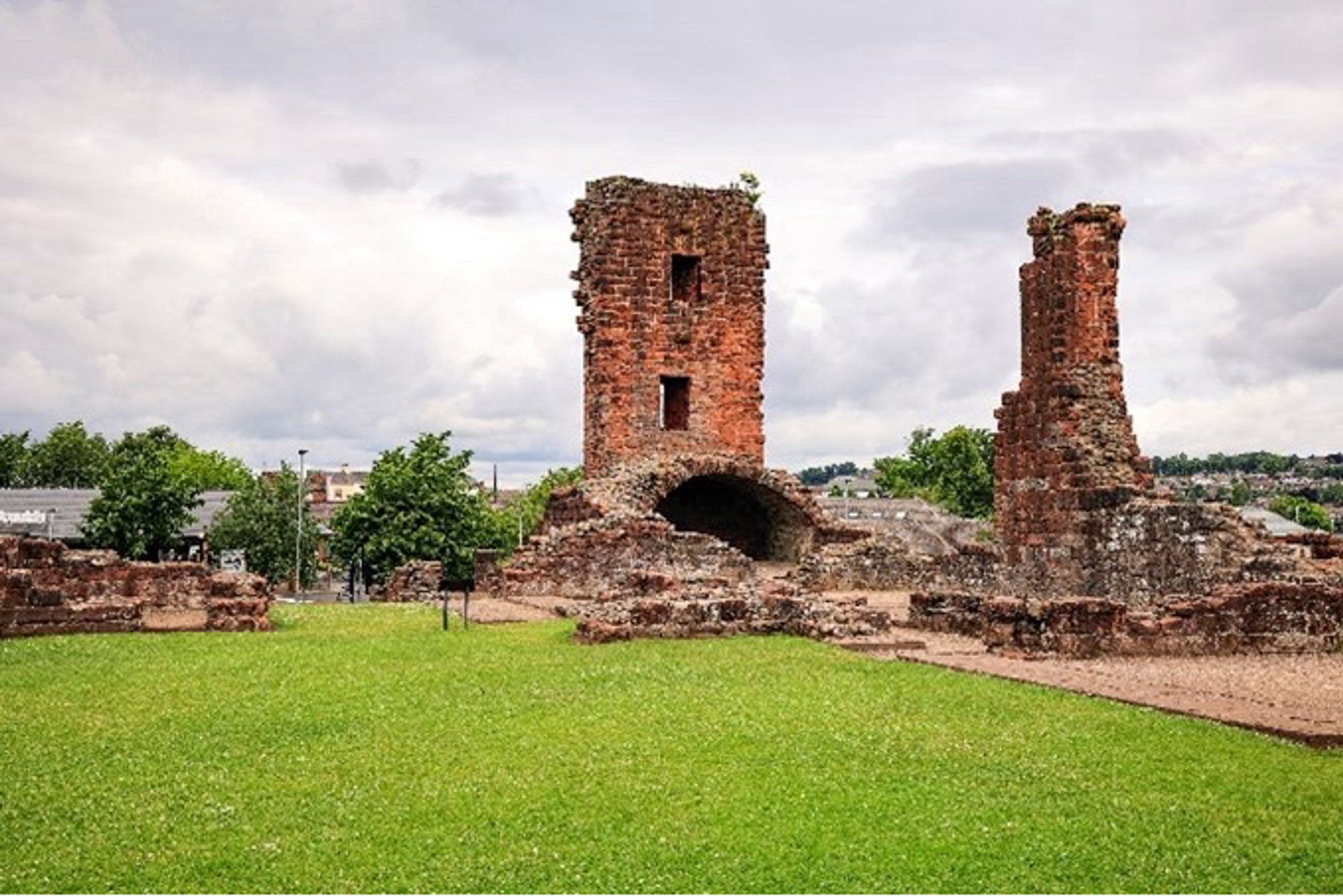 Photograph of Penrith Castle. There are few upstanding walls beyond shin height in this view; there are two tall stacks of stone with some flat faces which survive. The perspective of the photo is the same as the reconstruction.