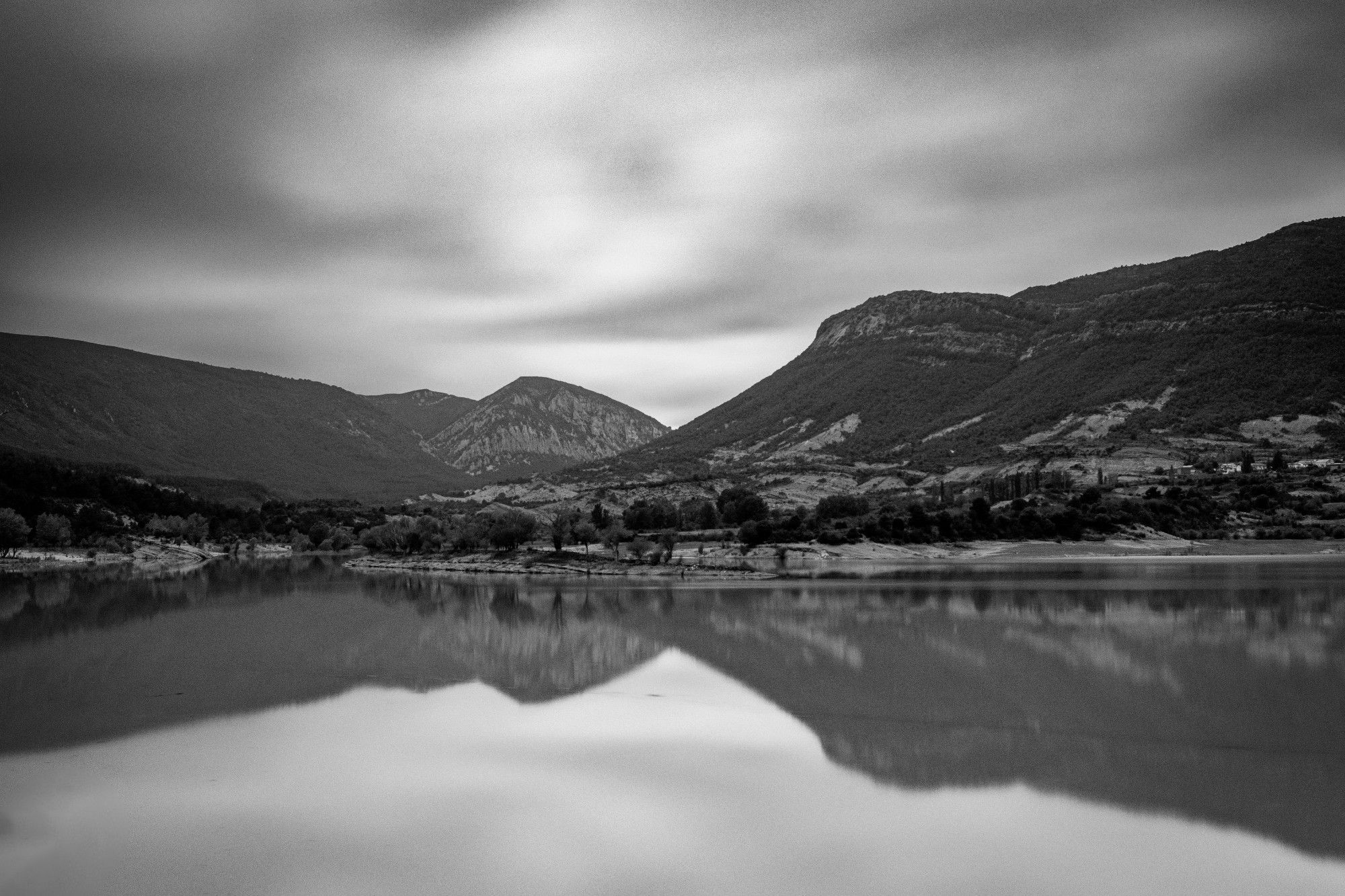 Vista en blanco y negro del pantano de Arguis (Huesca) en un día nublado. Se puede ver parte de la Sierra de Gratal reflejada en las aguas del pantano y en la parte superior algunas nubes en larga exposición.