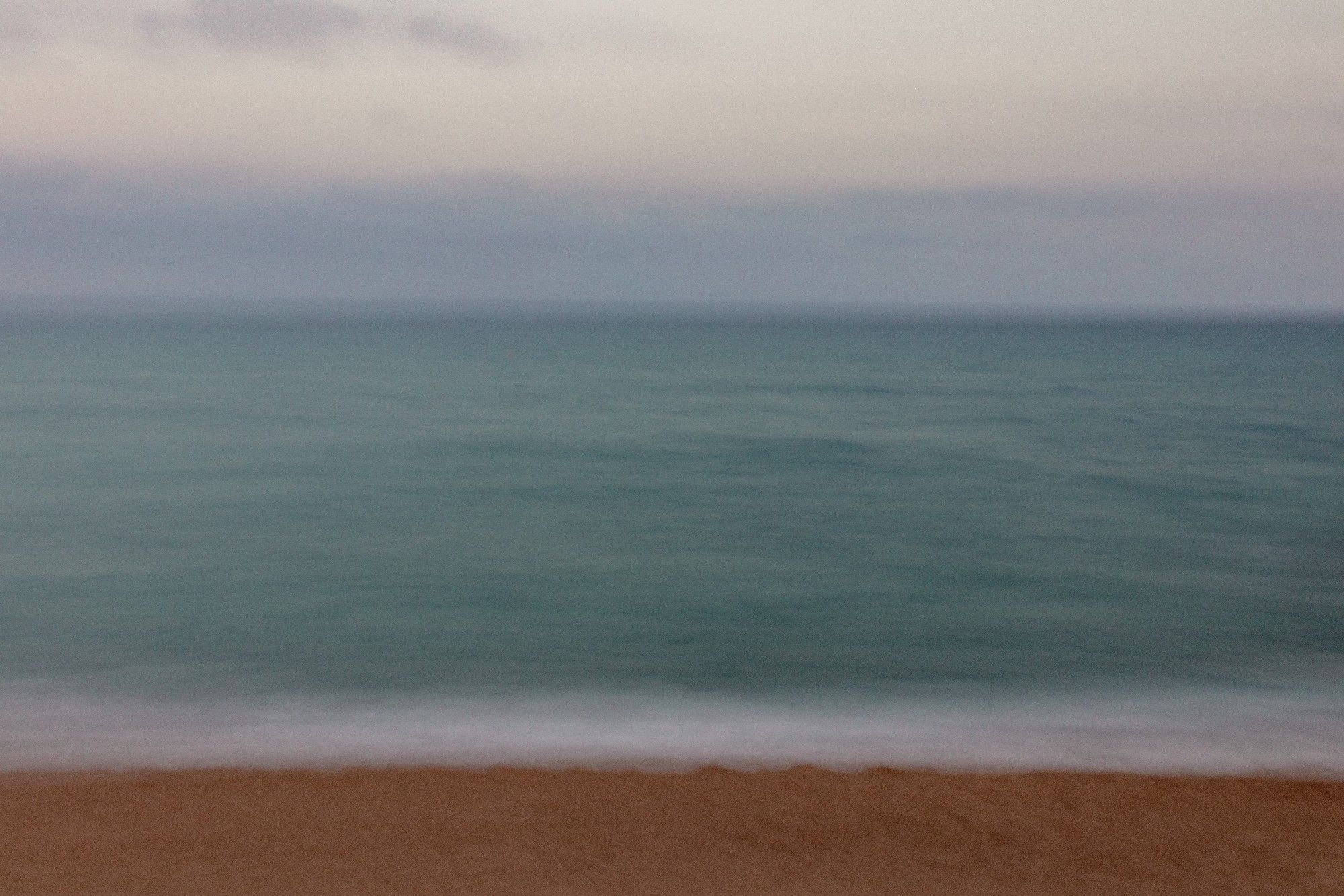 Fotografía usando larga exposición y moviendo ligeramente la cámara donde se ve el mar en una gran franja central y en la parte inferior, algo de tierra de la playa mientras en la superior un algo del cielo difuminado.