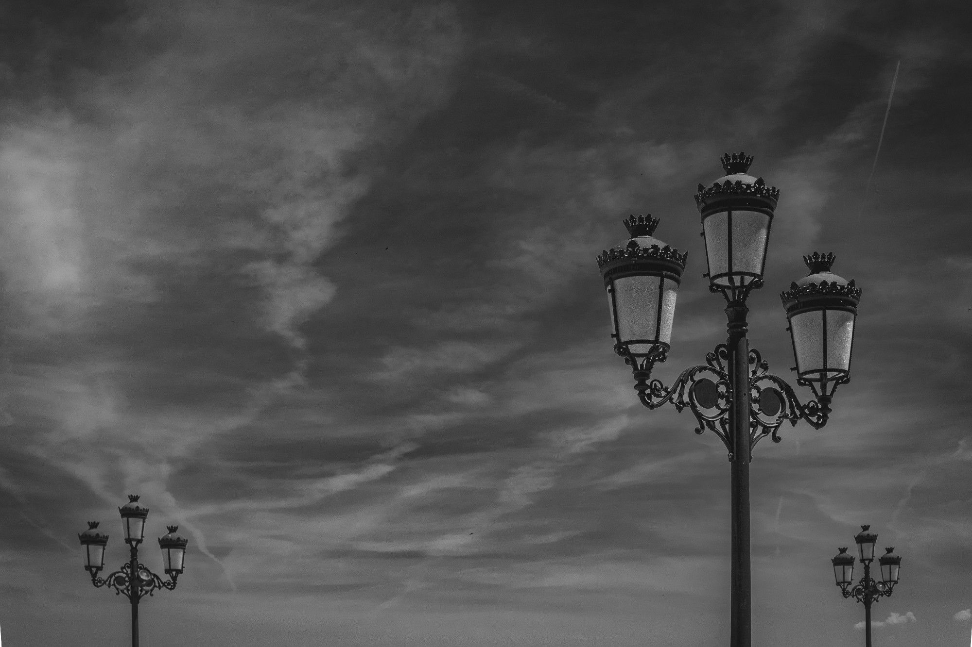 Fotografía en blanco y negro donde se ven tres farolas que a su vez cuentan con tres faroles.