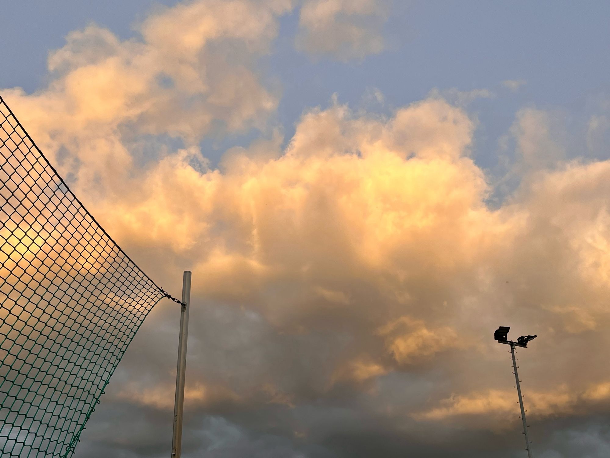 Wolkig-sonniger Abendhimmel. Links Fangnetz. Rechts Lichtmast