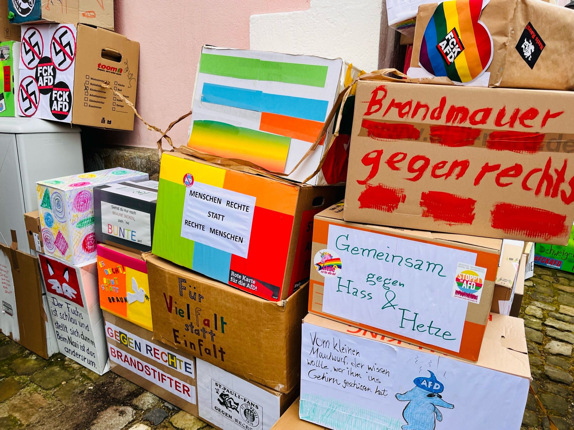 Bunte, mannshohe Mauer Aus Umzugskartons entlang einer Kirche auf Kopfsteinpflaster. Kartons mit vielen verschiedenen Anti AFD und Anti Nazi Sprüchen.