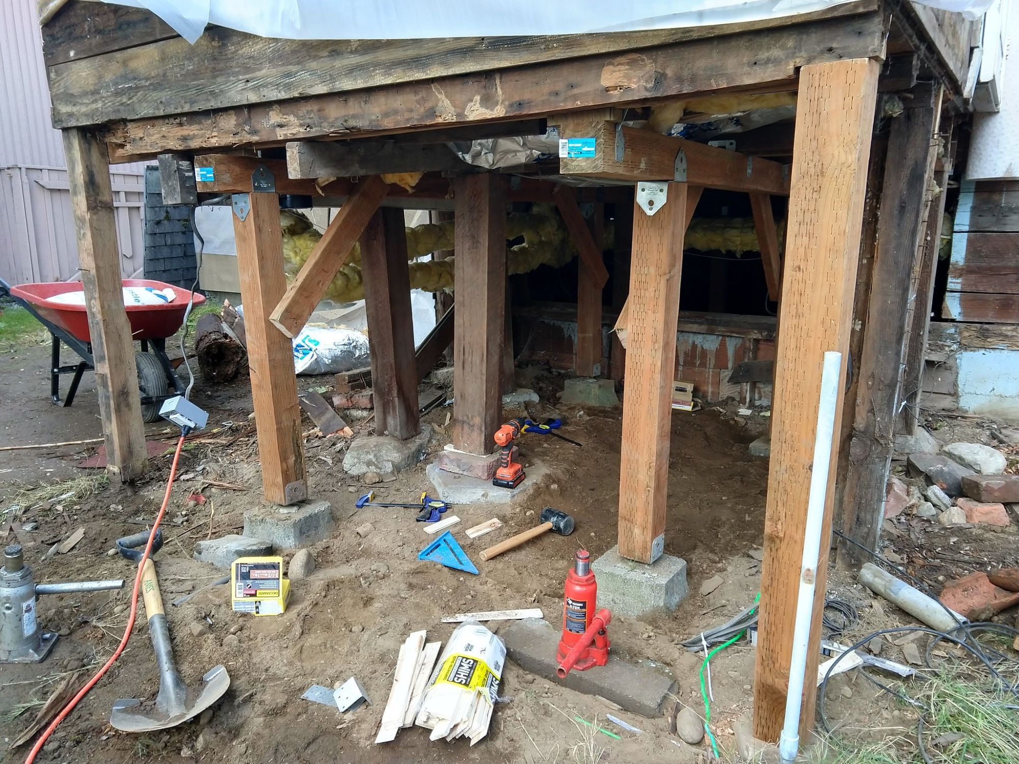 The support under a small building structure. While there are still several posts being supported by rocks and random bricks, they have been joined by 4x4s attached to deck blocks, with 4x4 beams supporting the joists and appropriately braced. One of the most rotten supports has been replaced by a fresh, pressure-treated 4x4 post.