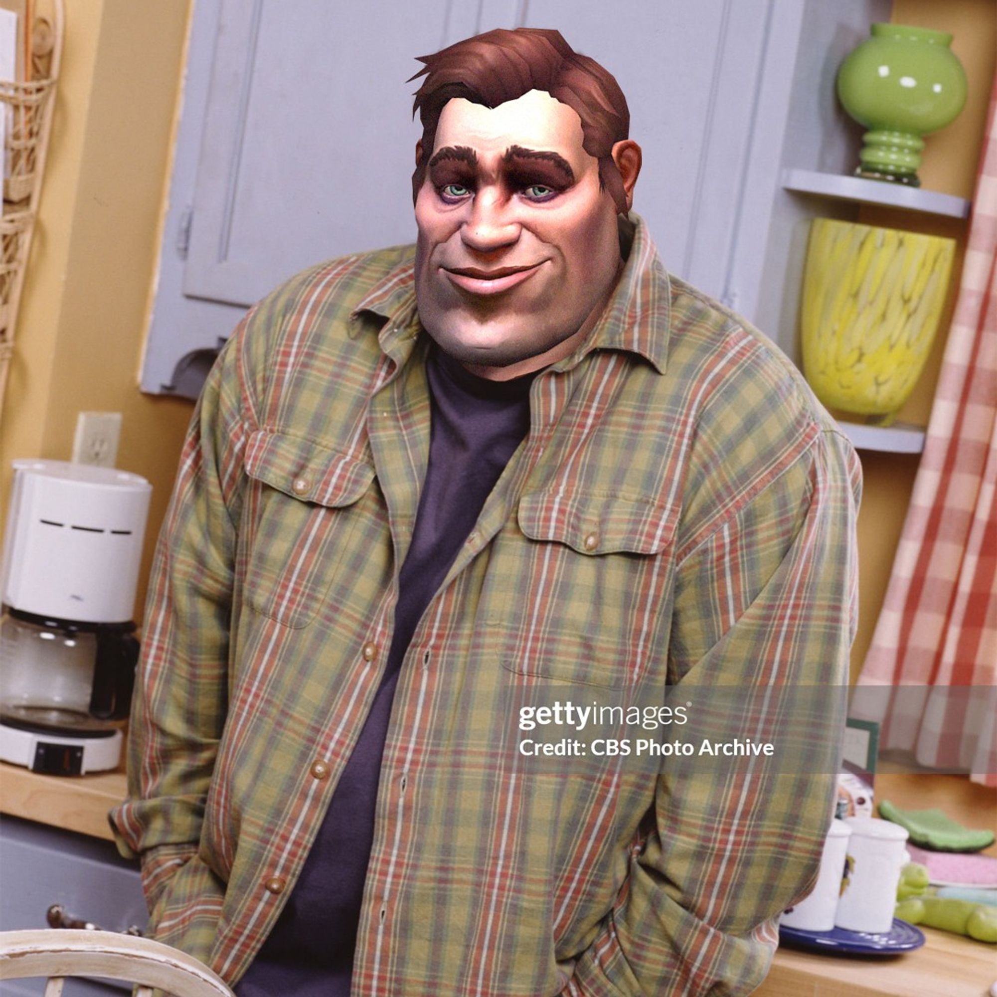 An image of actor Kevin James with his hands in his pockets and shrugging his shoulders, except the face of an in-game Kul Tiran character is superimposed over top. The Kul Tiran face is similar to the actor’s with short brown hair and a humble expression. He is wearing blue jeans and a purple t-shirt with green and red plaid flannel over top. A 90s sitcom kitchen fills the background with cream-colored walls, lavender cabinets, and a wooden counter with a coffee machine sitting to the side.