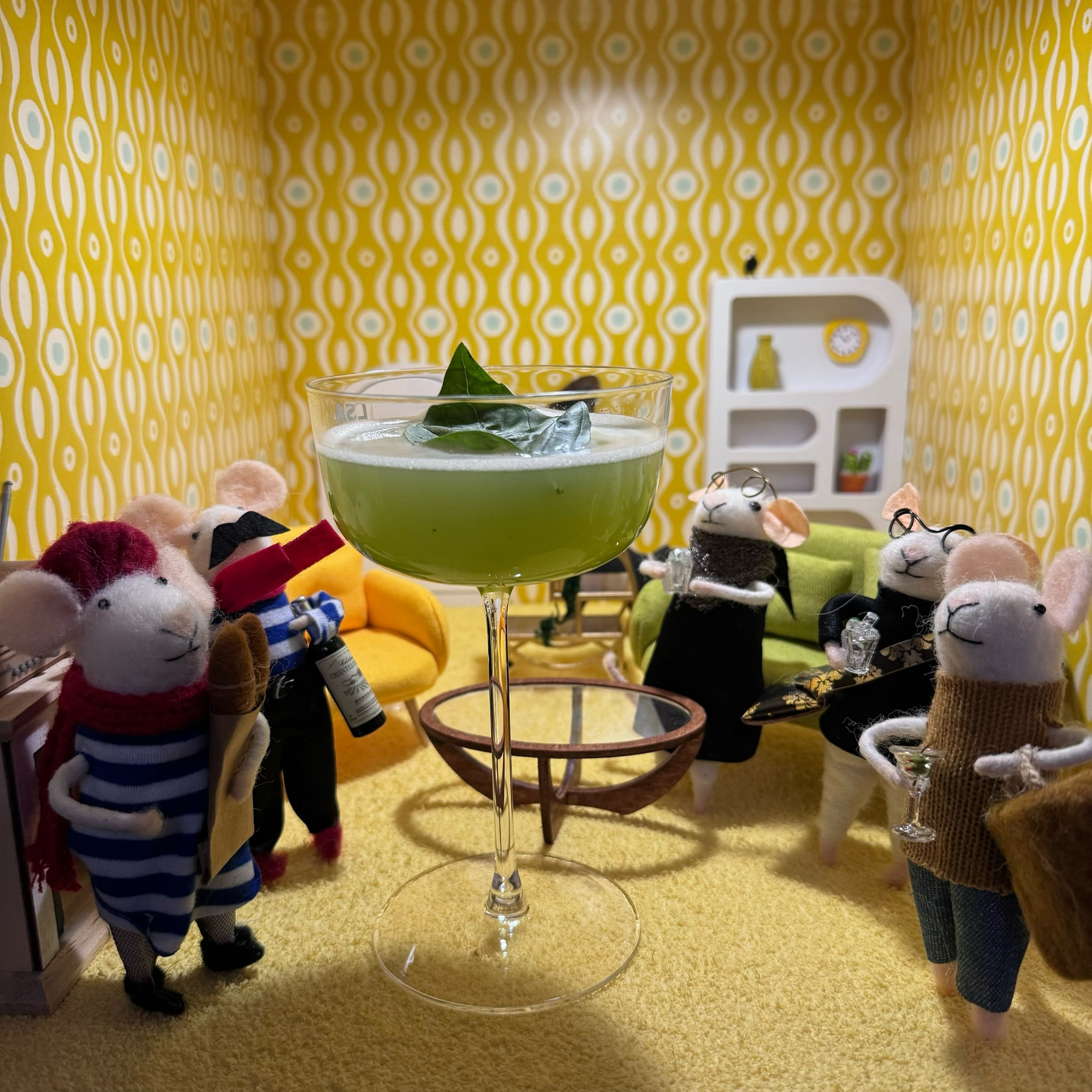 A large, green cocktail, garnished with a basil leaf. Five felt mice are gathered around the cocktail.