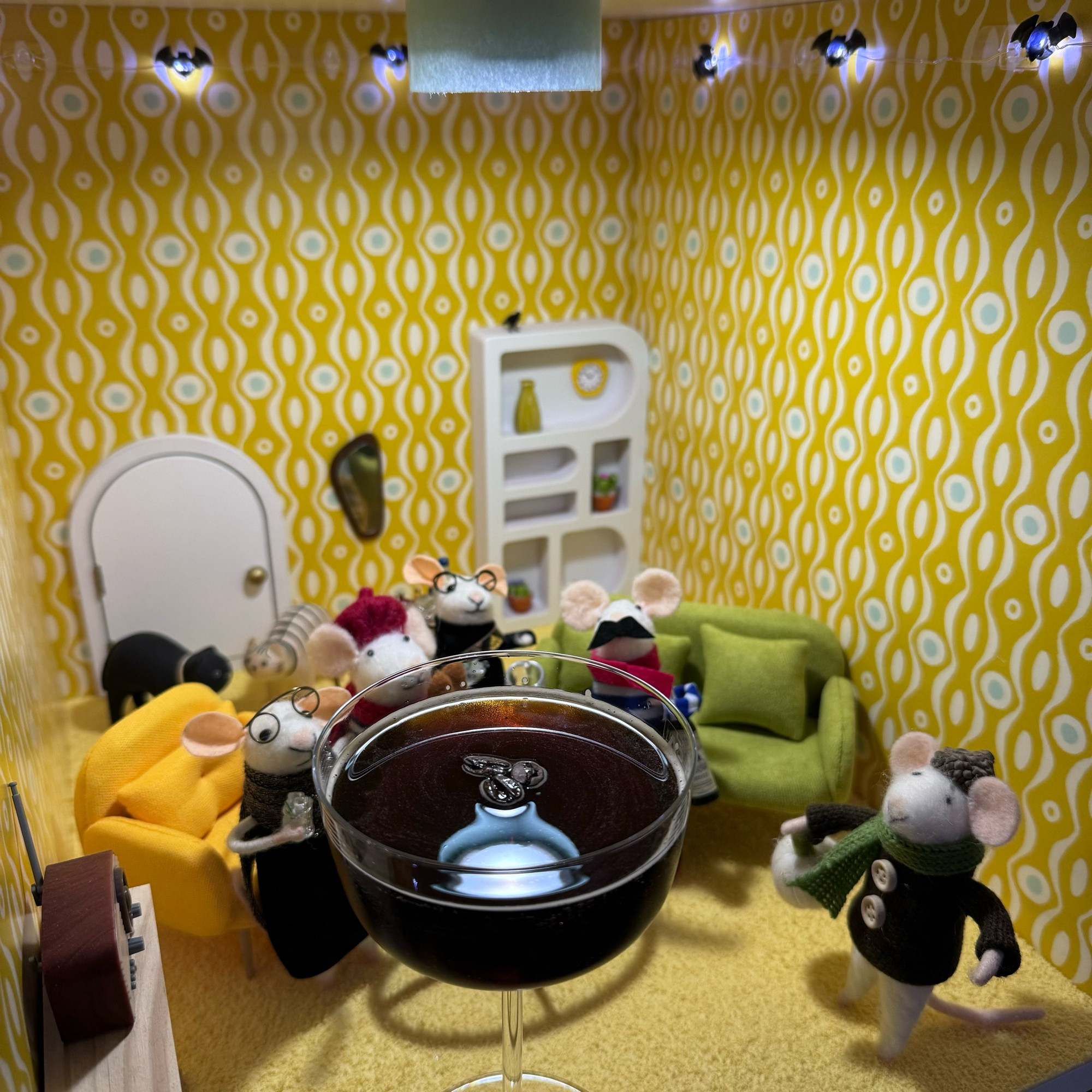 Five felt mice gathered around a giant espresso martini. Festive bat shaped lights are strung round the top of the walls of their living room.