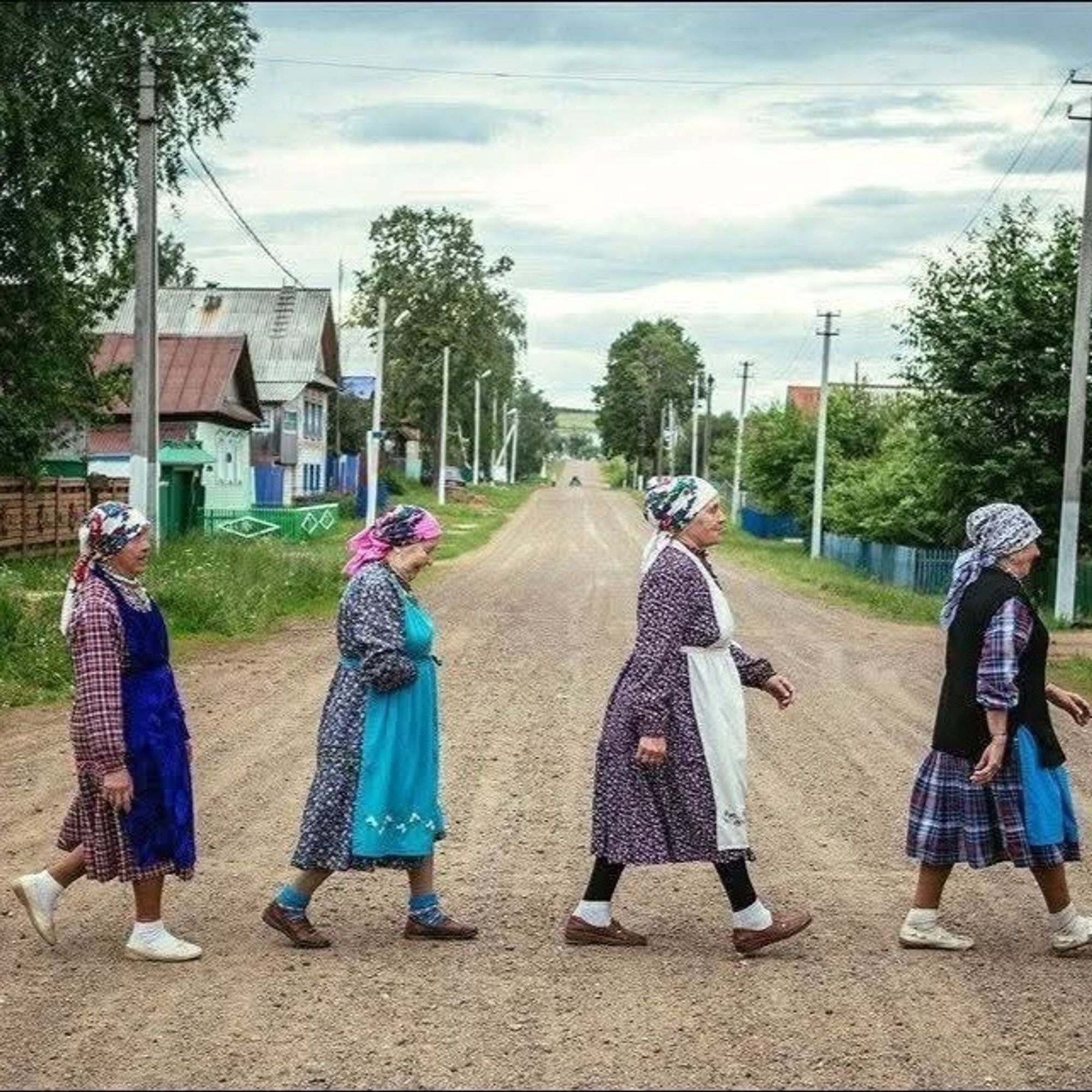 Vier sehr ländlich gekleidete, nicht mehr ganz junge Frauen mit bunten Kopftüchern, Kittelschürzen und grobem Schuhwerk überqueren im Gänsemarsch eine dörflichen Schotterstraße in Osteuropa.