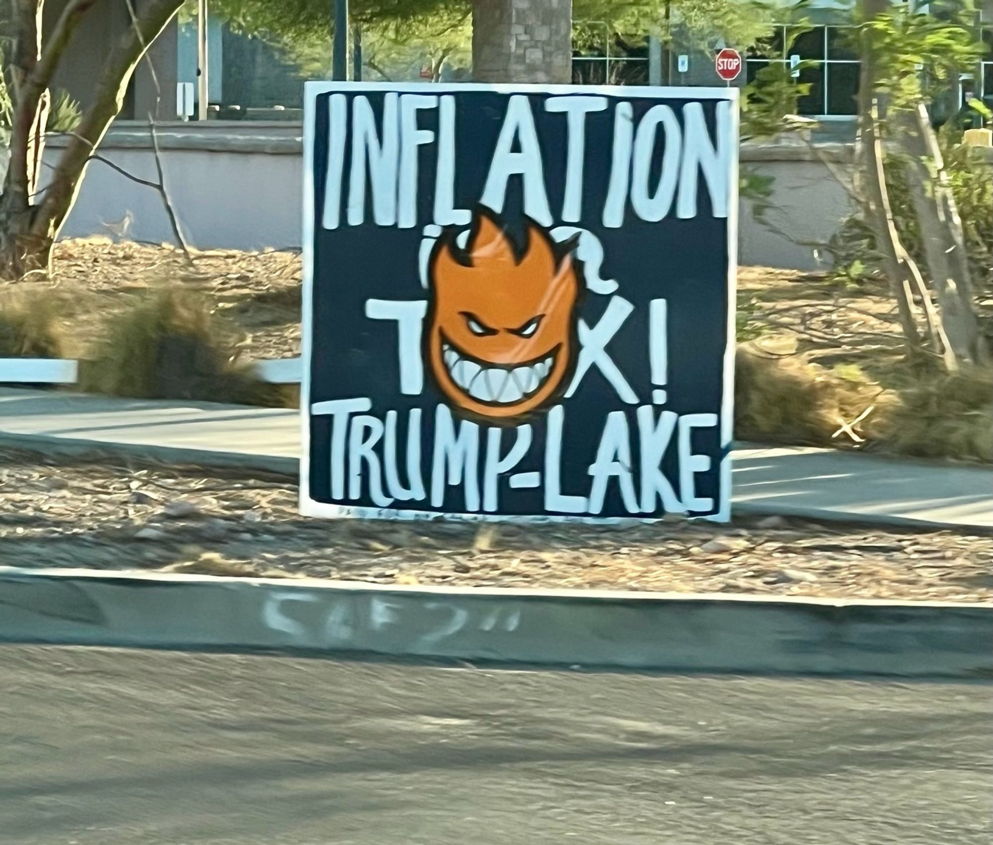 A DIY sign along the road that’s just plain black paint with white text reading “inflation is a tax! Trump-Lake”

someone has placed a large orange Spitefire smiling flame logo in the dead center, covering half the text.