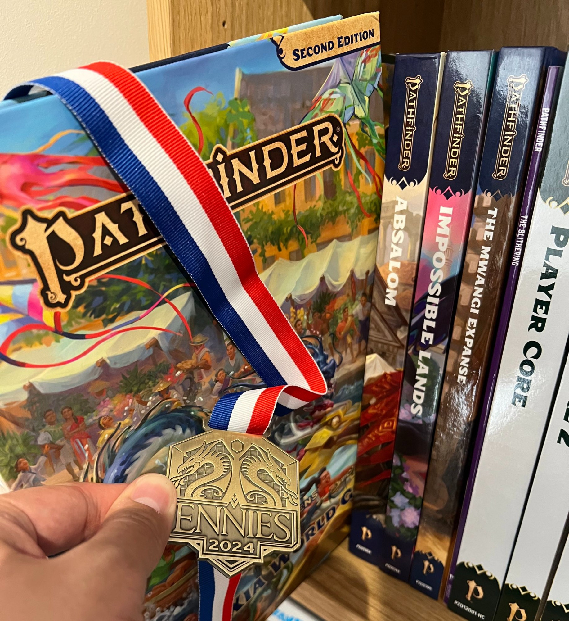 Me holding up my 2024 ENNIE medal in front of a shelf of Pathfinder books.