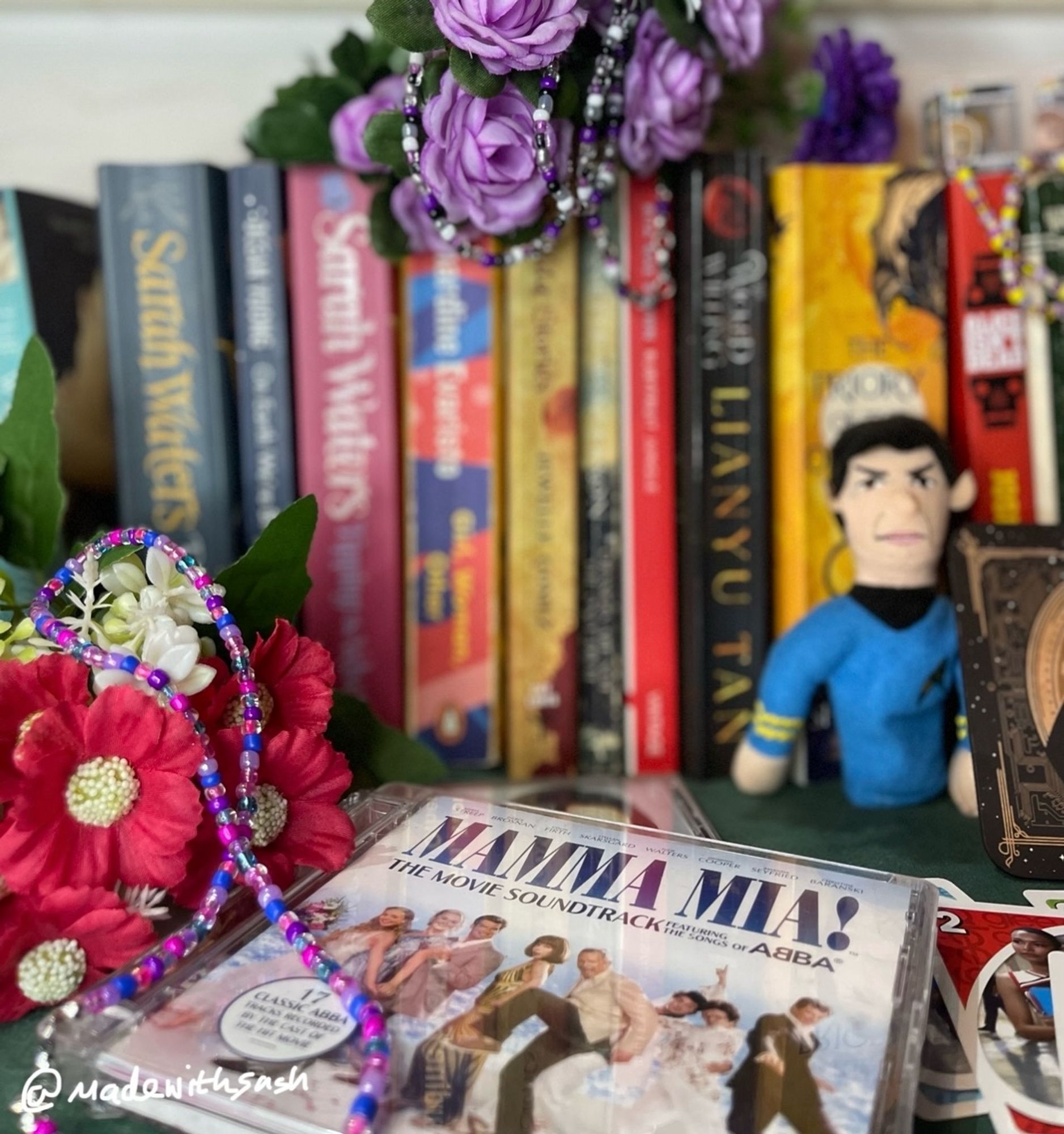 set up showing off some of our queer pieces of jewellery so far. the set up is a bunch of queer books in the back, lots of random fake flowers, a spock finger puppet, the mamma mia soundtrack cd , a TNG coaster of data as sherlock. on the left is a bisexual choker: pinks, purples, blues, resting on the cd and red flowers. in the back is an ace choker: blacks, whites, purples, greys, hanging on top of the books off purple flowers. our insta @madewithsash in left corner