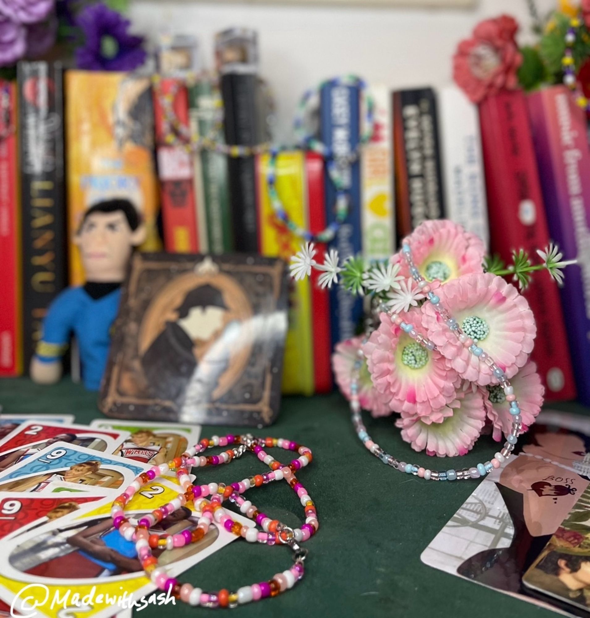 same set up just moving along to the right. spock finger puppet is on the left with data coaster on his right. there is a lesbian choker: lots of pinks, whites, oranges, resting on top santana and brittany glee uno cards lol. a trans choker: whites, pinks, blues, resting around pink flowers, dr frankenfurter bookmark to the right. in the back but blurry is a gay choker: blues, whites, greens and intersex: purples and yellows. our insta at @madewithsash left corner