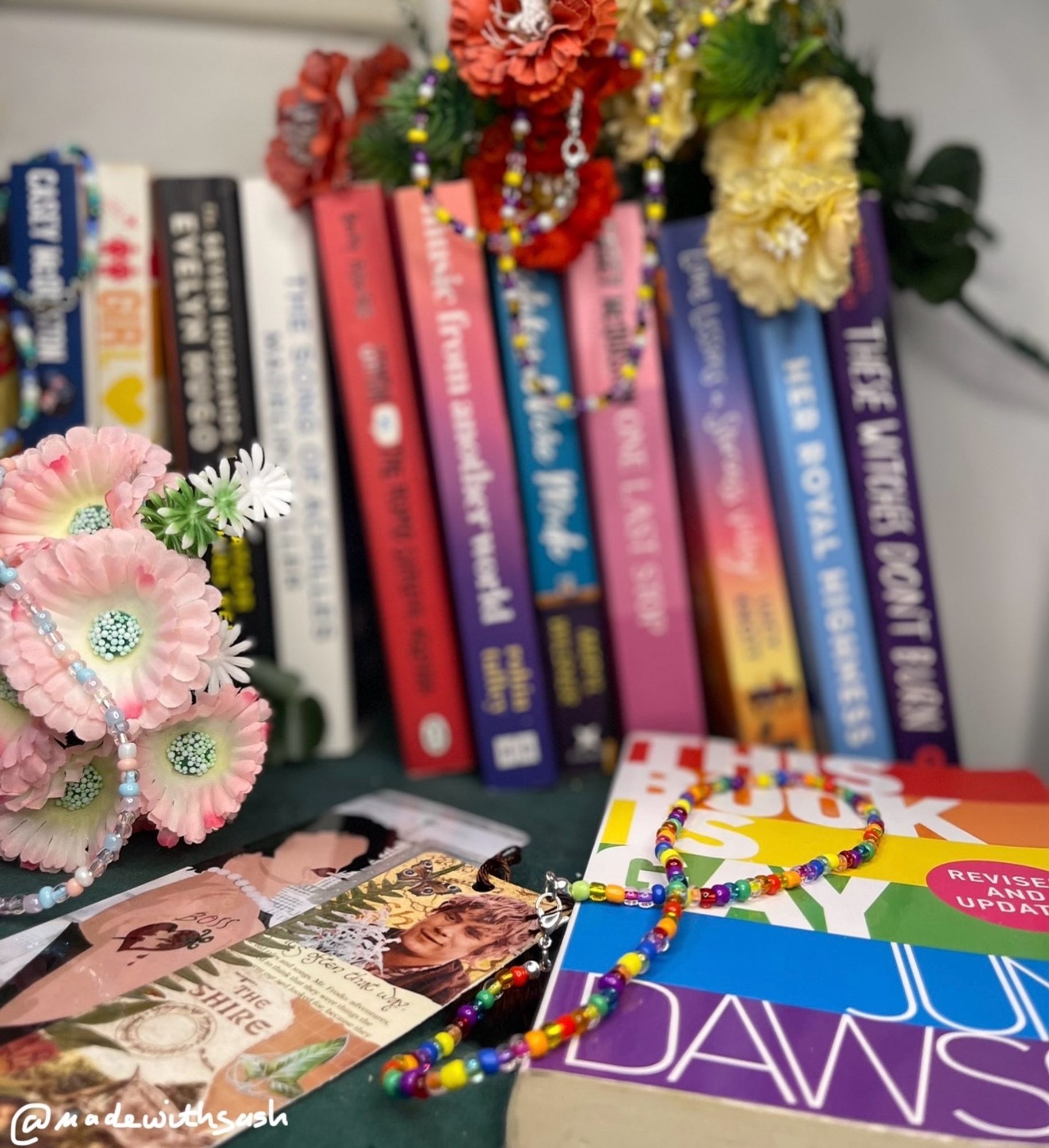 moving along to the right at the end. a queer choker mirroring the 6 colour flag resting on “this book is gay” by juno dawson with a sam/frodo bookmark to the left and the frankenfurter one on sam’s left. in the back but blurry is a nonbinary choker: whites, yellows, purples, blacks, hanging from red and yellow flowers ontop of the books. our insta @madewithsash left corner