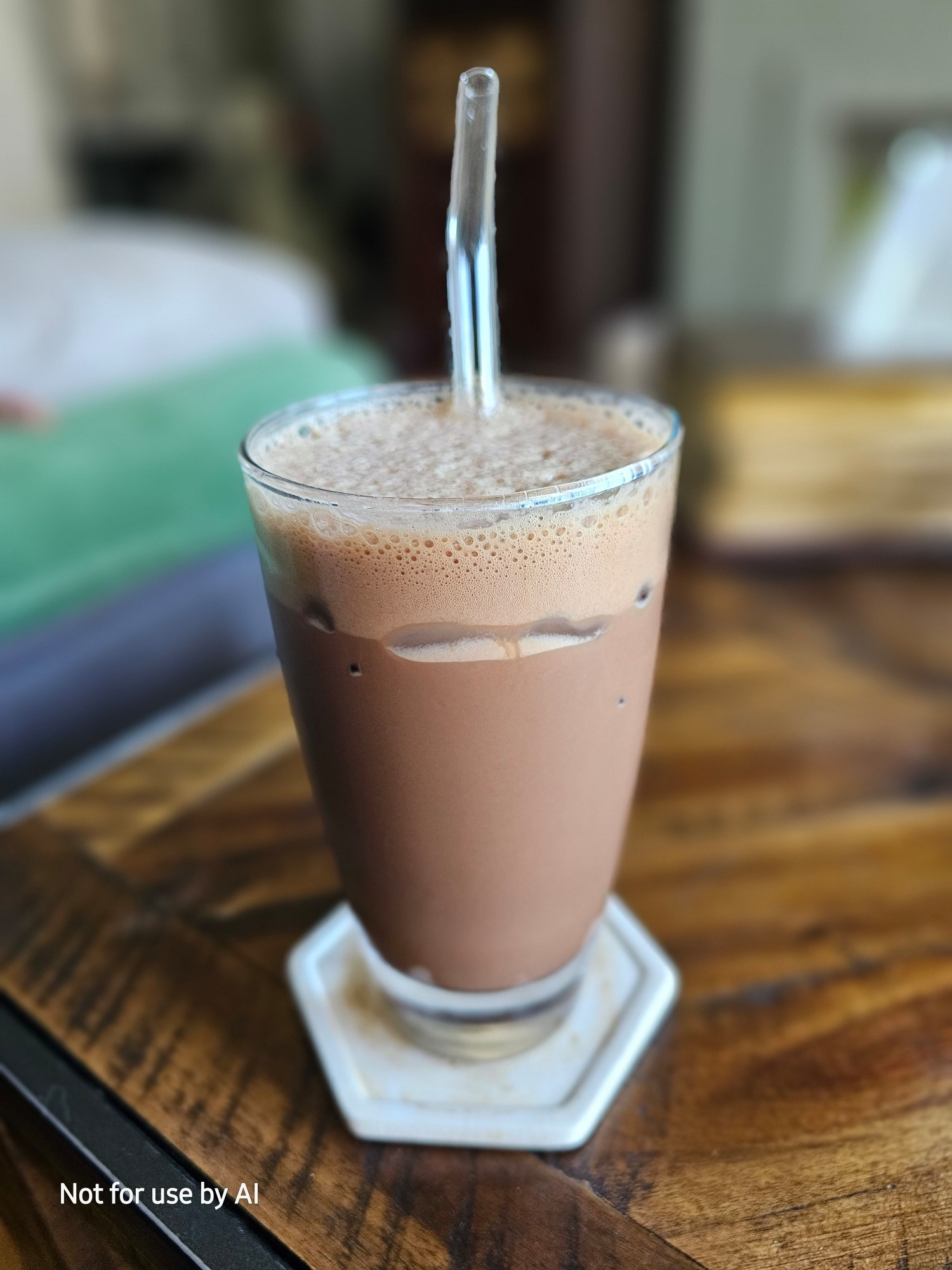 A clear glass on an off-white coaster, with a dark chocolate, coconut, iced mocha, and a glass straw, in it. There is a watermark in the lower lefthand corner that reads, "Not for use by AI".