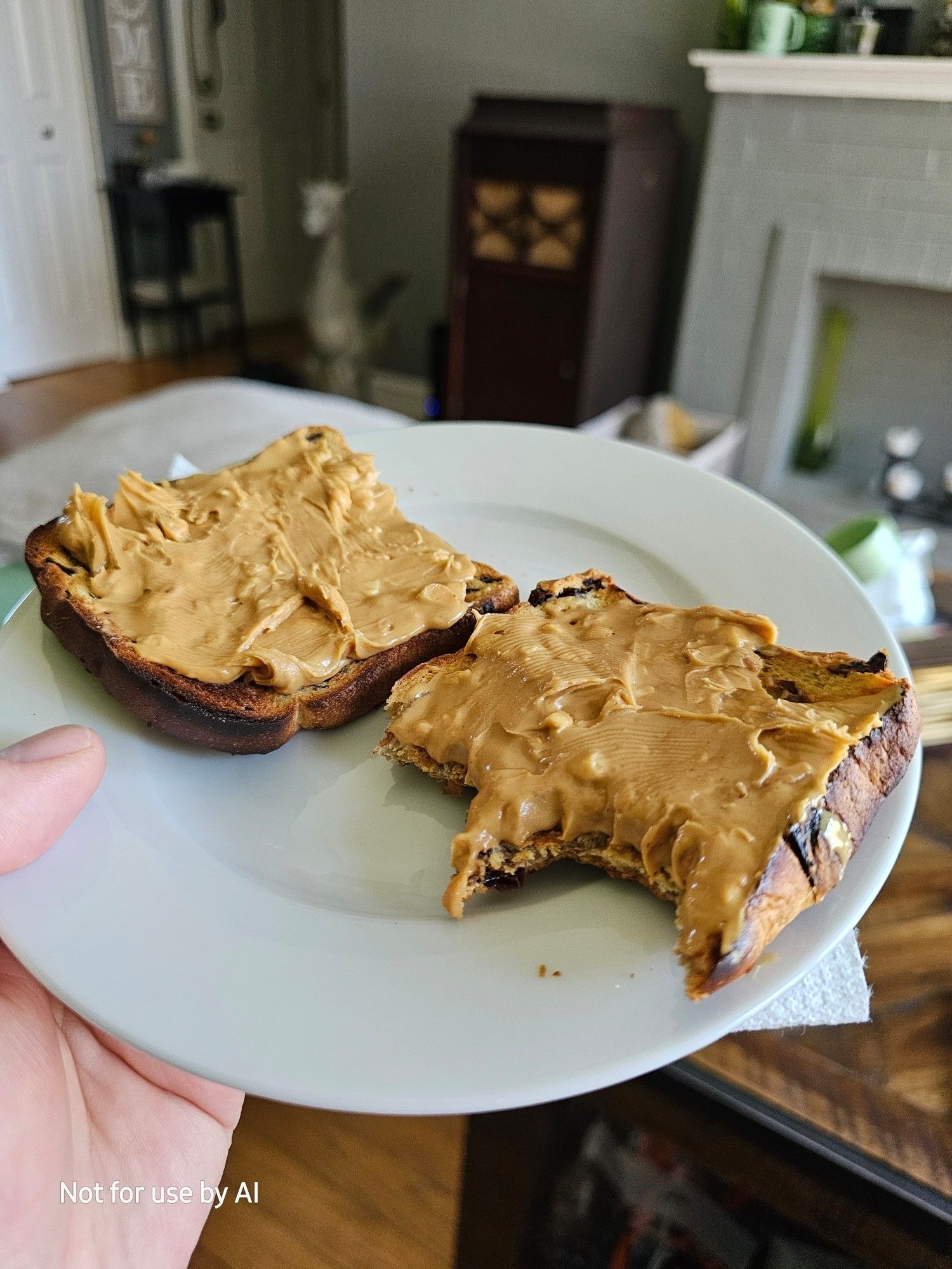 Two pieces of cinnamon raisin toast, one with two bites taken out if it, slathered with crunchy peanut butter, on a white plate. There is a watermark in the lower lefthand corner that reads, "Not for use by AI".