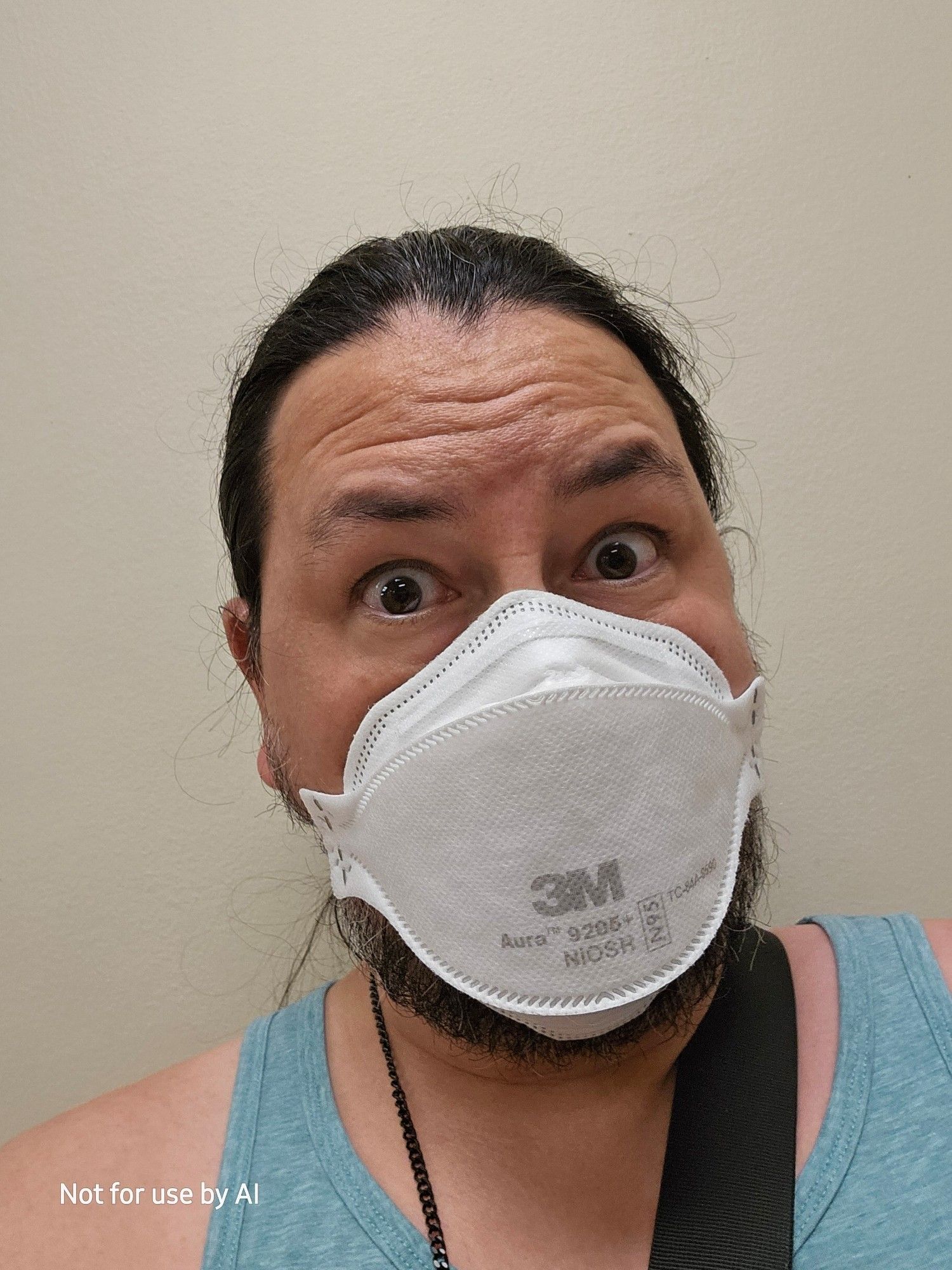 A picture of me, a 46-year-old, queer, cisgender, biracial (Japanese | Anglo European) male human with chocolate brown eyes, pepper-and-salt hair, & salt-and-pepper facial hair. My hair is pulled back in a rushed, disheveled ponytail. I'm wearing a white, 3M N95 mask, and a turquoise tank top. My black, metal glasses chain and the black strap to my purse slung across my body are visible. I'm looking wide-eyed at the camera, head cocked slightly to the right. There is a watermark in the lower lefthand corner that reads, "Not for use by AI".