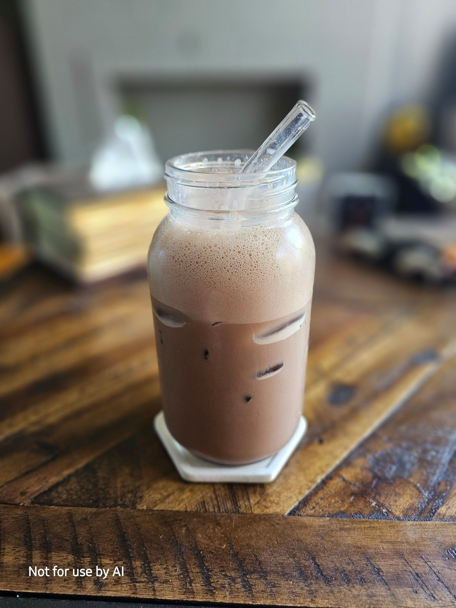 A medium-to-large mason jar, with an iced, coconut, dark chocolate mocha, and a glass straw in it. There is a watermark in the lower lefthand corner that reads, "Not for use by AI".