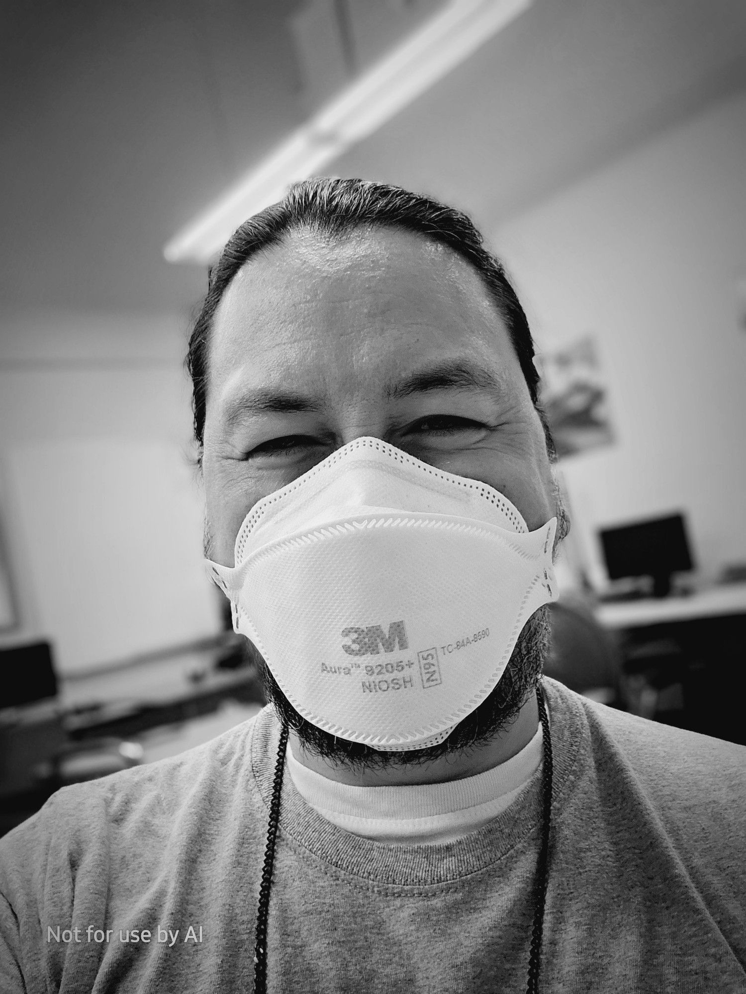 A black-and-white, vignette-shaded picture of aA picture of me, a 46-year-old, queer, cisgender, biracial (Japanese | Anglo European) male human with chocolate brown eyes, pepper-and-salt hair, & salt-and-pepper facial hair. I am wearing a white 3M N95 mask, a gray t-shirt, and a white undershirt. My black, metal glasses chain is visible. My hair is pulled back in a ponytail, and I'm smiling at the camera. There is a watermark in the lower lefthand corner that reads, "Not for use by AI".