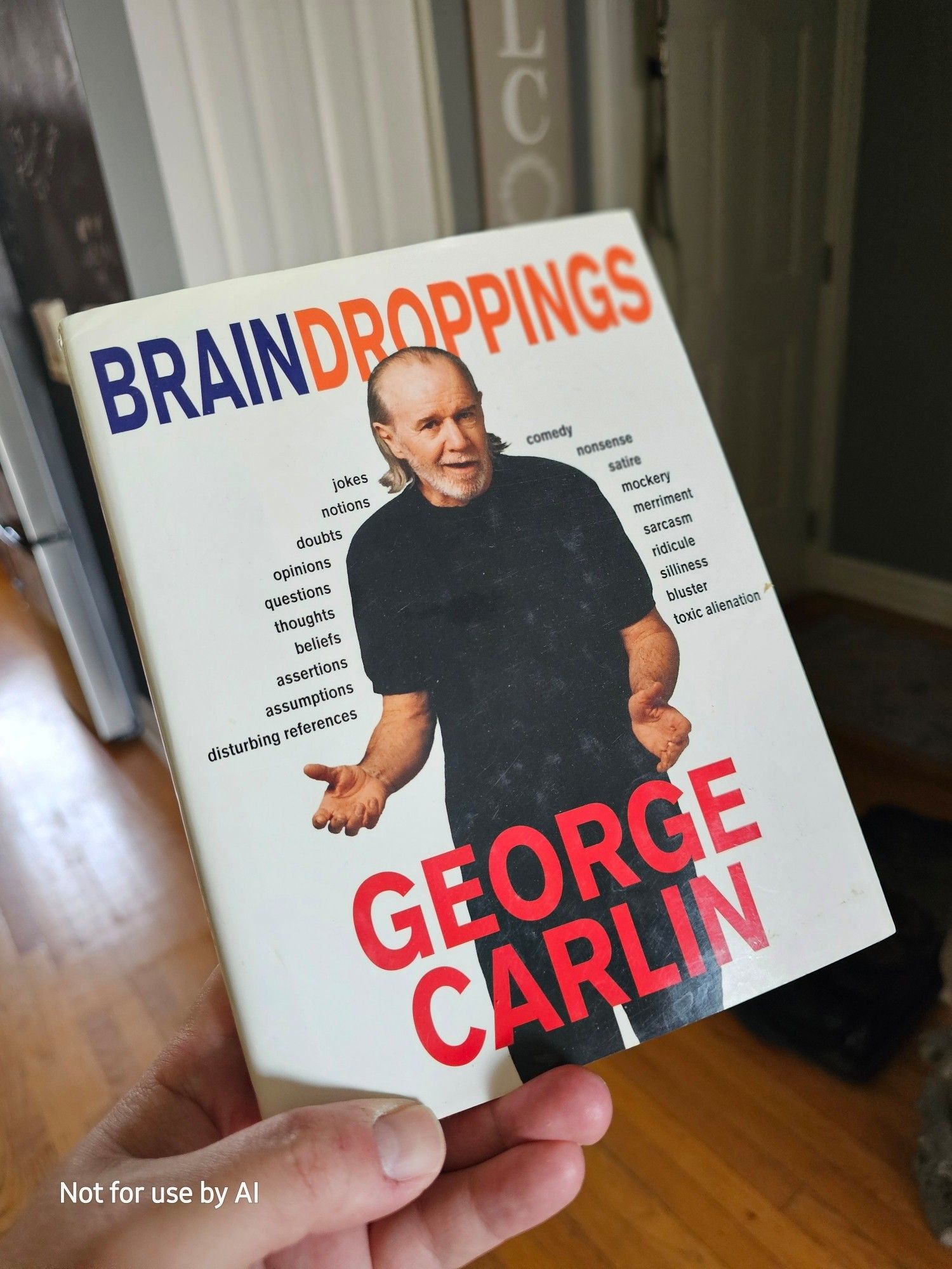 Me holding a hardcover copy of George Carlin's book, "Brain Droppings". There is a watermark in the lower lefthand corner that reads, "Not for use by AI".