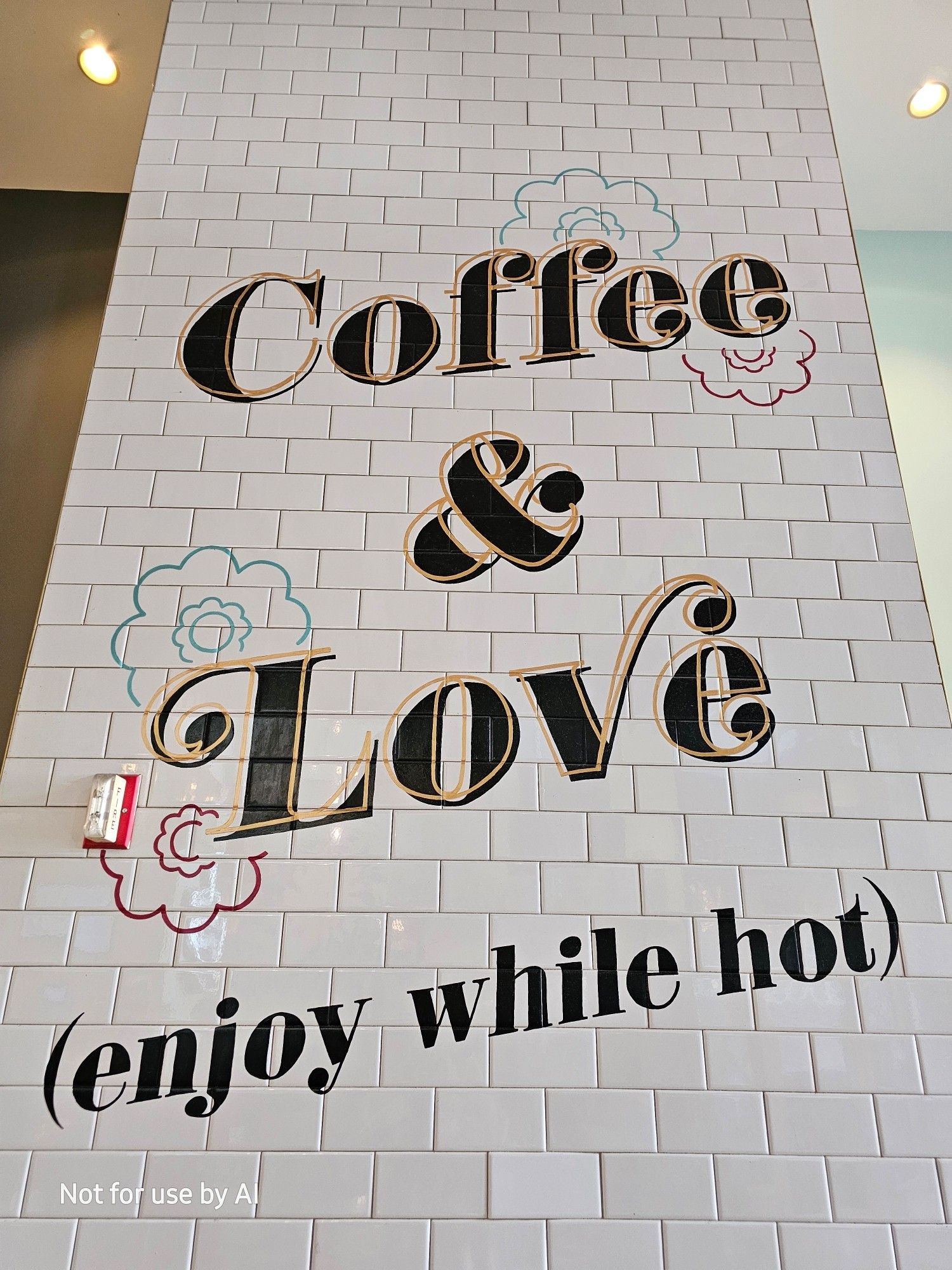 A white, shiny brick wall, printed with an angled, black print that reads, "Coffee and Love (enjoy while hot)". There are outlines of flowers around the "e" of "Coffee", and the "L" of "Love". There is a watermark in the lower lefthand corner that reads, "Not for use by AI".