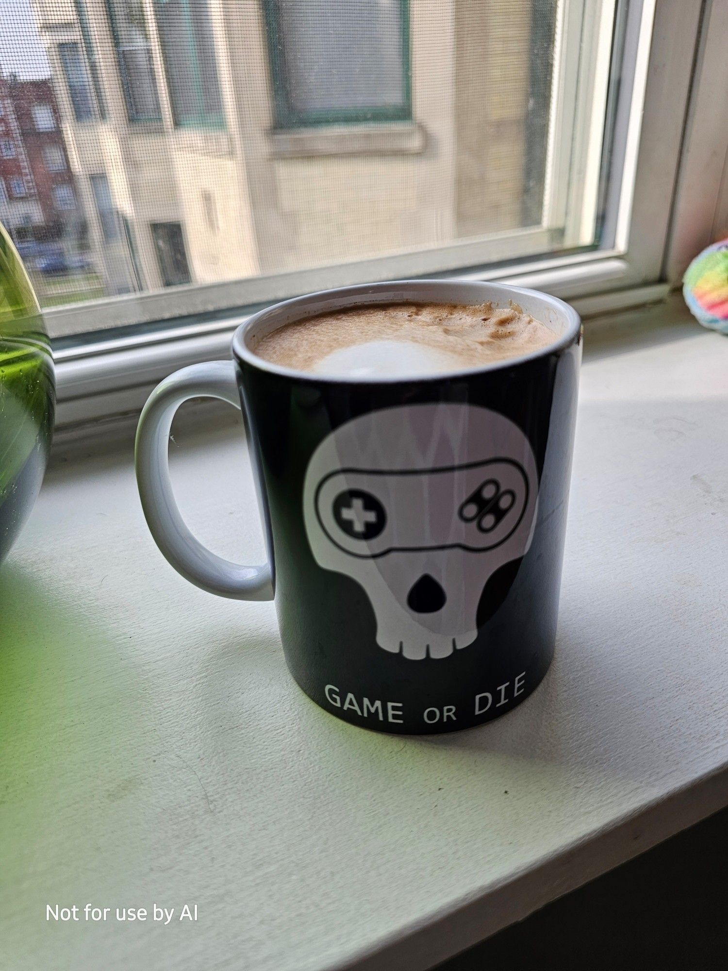 A white mug, overlaid with a black skin, allowing for the visual of a stylized white skull, missing its jawbone, with four rounded teeth, and a stylized game controller reminiscent of a Super Nintendo controller where it's eyes would be; the D Pad is its right eye, and the controller buttons are its left eye. At the bottom of the mug, also in white, are the words, all in caps, "GAME OR DIE". The word, "OR", is slightly smaller than the words on either side of it. The mug is sitting on our living room windows sill, and contains a homemade raspberry dark chocolate mocha. There is a watermark in the lower lefthand corner that reads, "Not for use by AI".