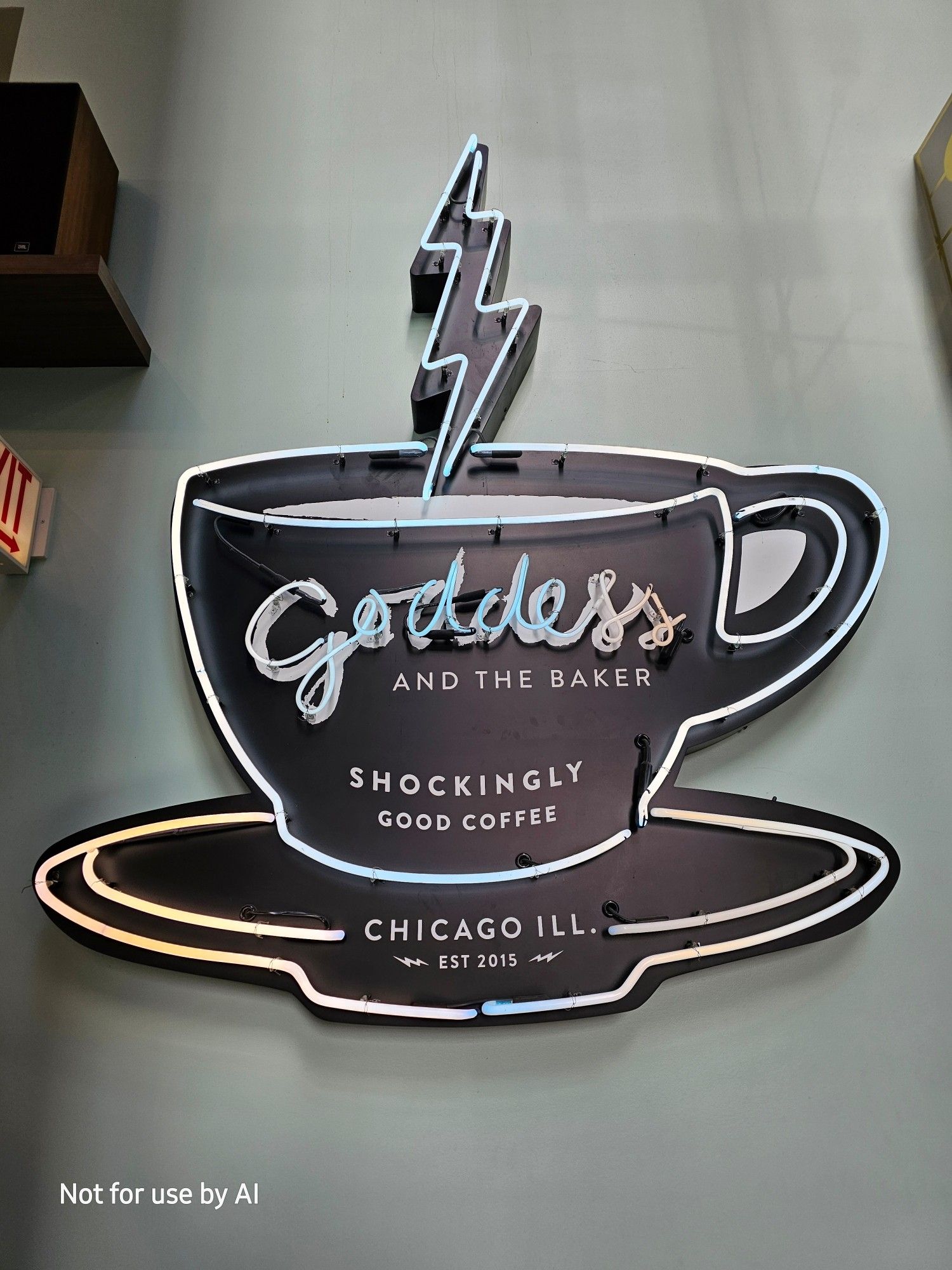 A large sign consisting of the logo for Goddess and the Baker, which is a black silhouette of a cup and saucer with a lightning bolt on place of the steam from the hot beverage, with "Goddess" written in cursive, and "and the Baker" in capitalized print. At the bottom of the cup, curved to align with the bottom of the cup, it reads, "shockingly good coffee". On the saucer, it reads, "Chicago Ill.", and under that it reads, "est. 2015", flanked by a pair of lightning bolts. The whole thing is outlined in neon lighting. "Goddess" is also in neon. There is a watermark in the lower lefthand corner that reads, "Not for use by AI".