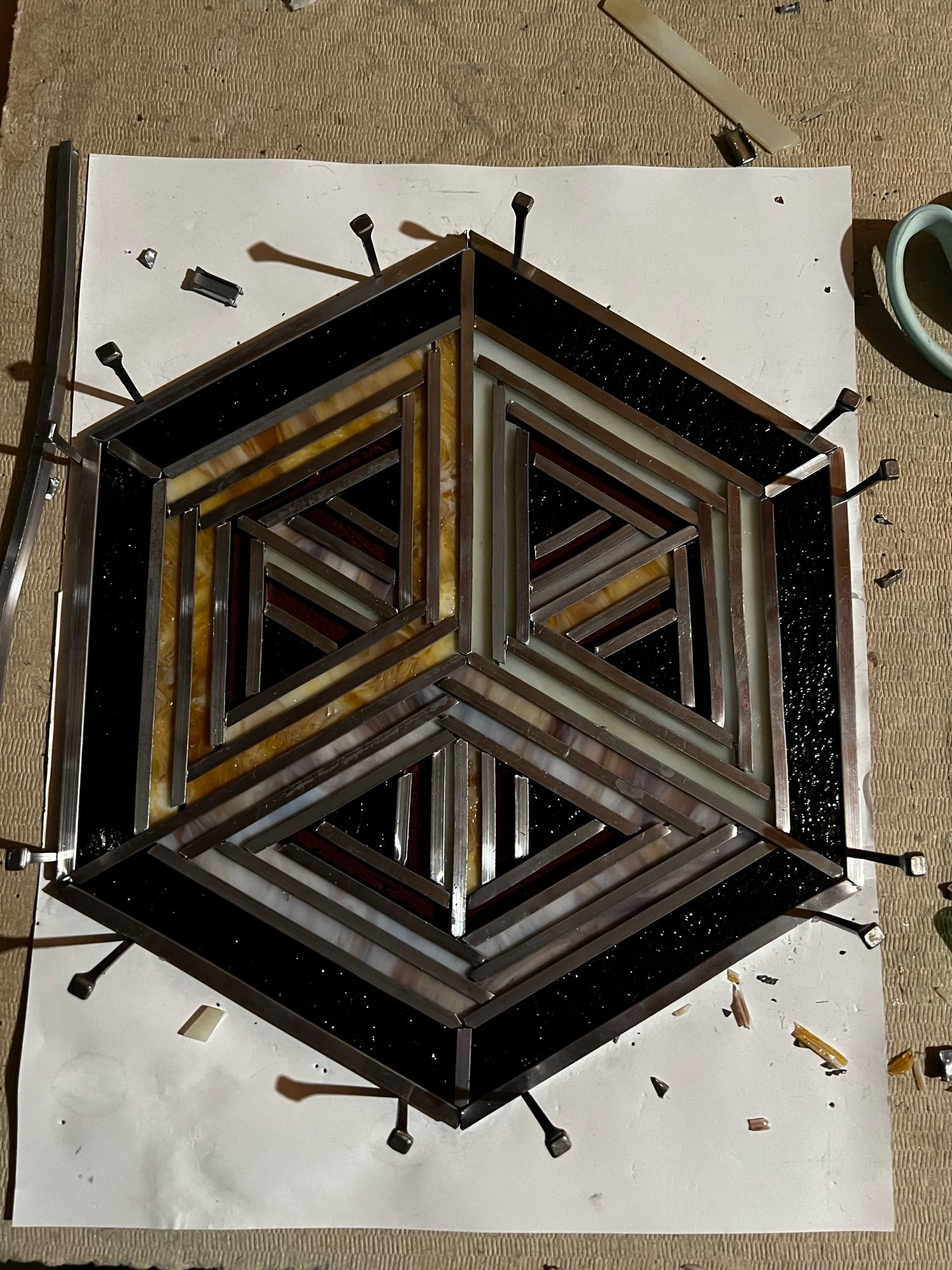 A hexagonal piece of stained glass still under assembly on a work surface, viewed from above. It's stylized to show the orthogonal projection of a cube's outline in three colors; gold, white, and purple. It's floating on a dark textured background. The line work is straight pieces of metal between each piece of glass, and the perimeter is bordered with horseshoe nails to help the piece hold its shape.