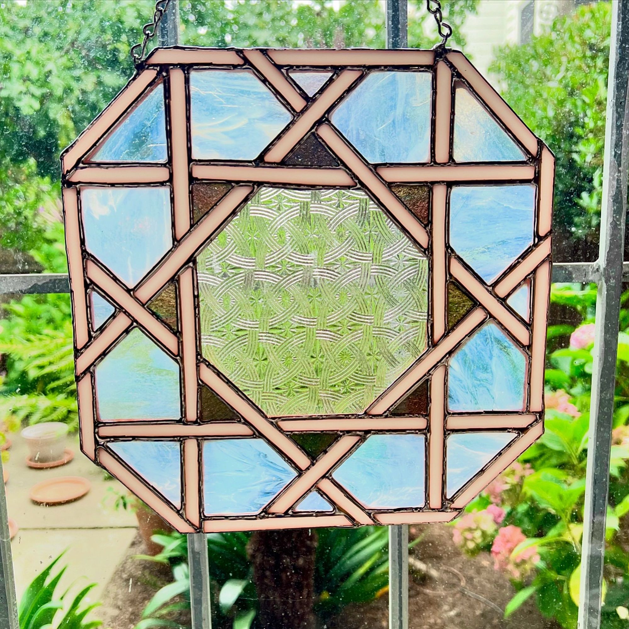 An octagonal stained glass piece, largely formed by a criss-crossing lattice of white ribbons, covering a field of milky white clear glass, at the center forming an octagonal sunburst with the points being made of multicolored glass, and the center being a textured transparent glass with interlocking rings and stars, hanging in front of an out-of-focus view of a garden