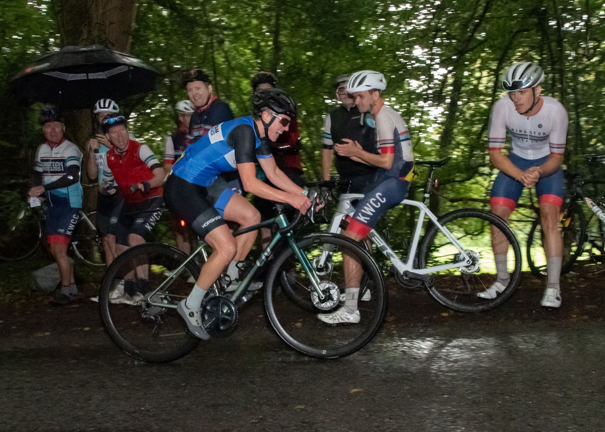 A woman cycles uphill. Other fellow cyclist cheer from the roadside, in the background. Rainy day, dark under the trees. Wet.