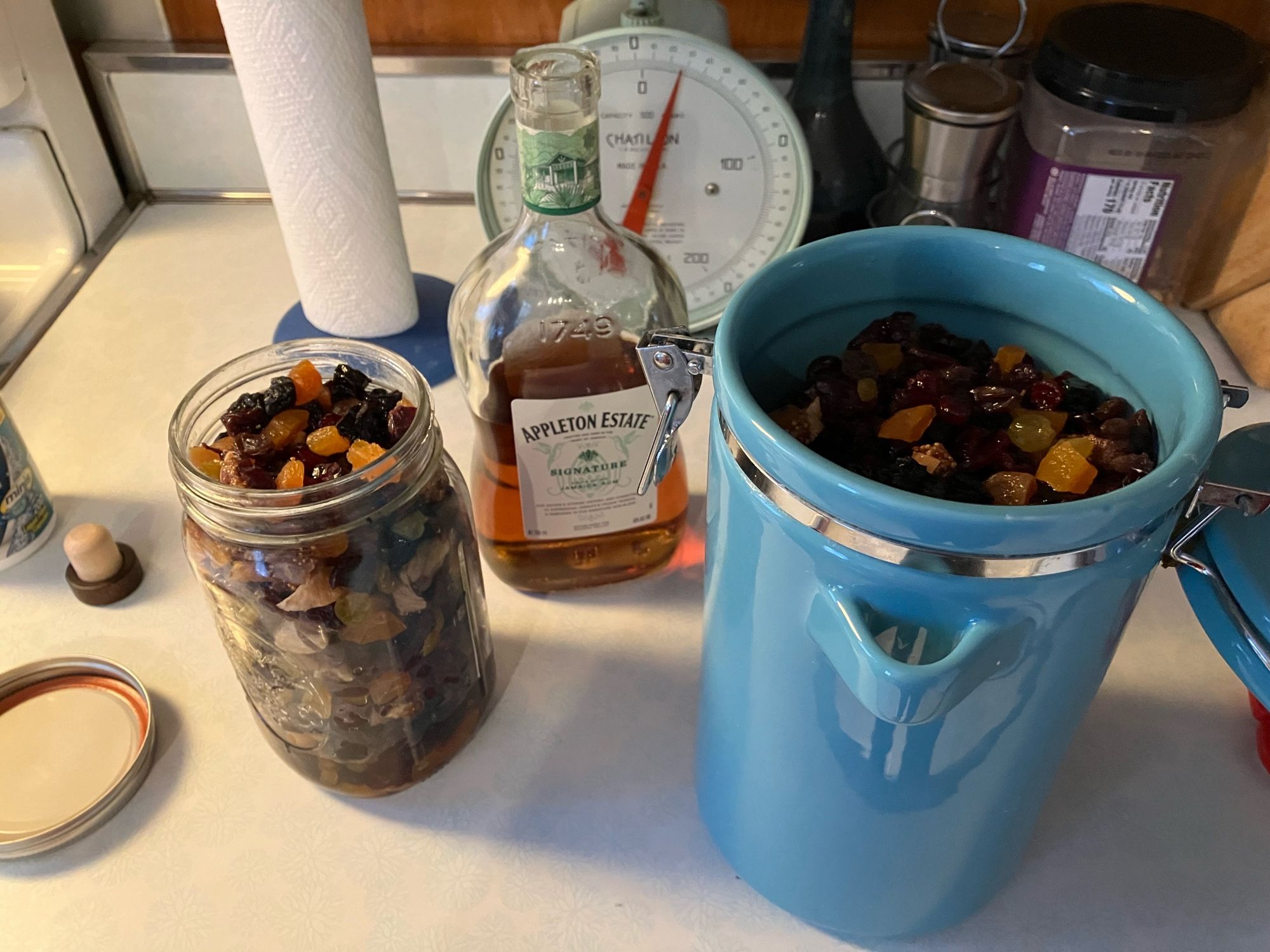 Fruit divided and now soaking in rum in a large glass jar and a large blue ceramic container. The Bottle of Rum is now empty.