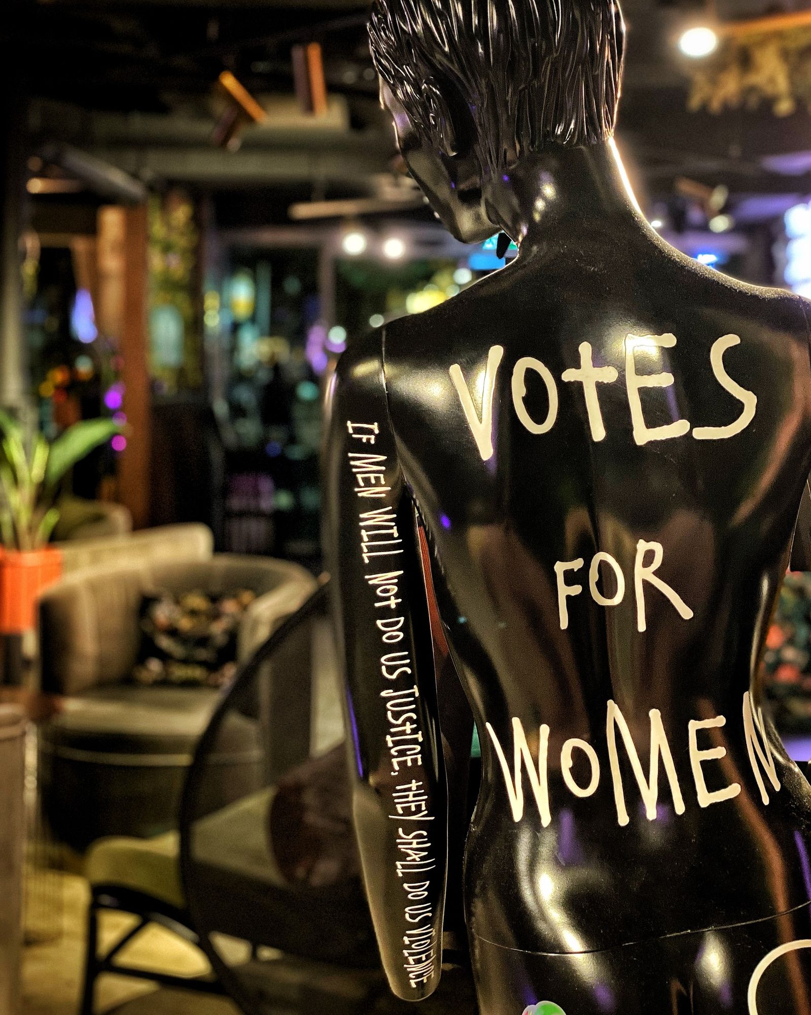 A black lifesize figurine of a woman is shown from behind. White lettering on her back reads “Votes for women”. Her left arm bears the text “If men will not do us justice, they shall do us violence”. In the background there are some comfy chairs, plants in a hotel lobby.