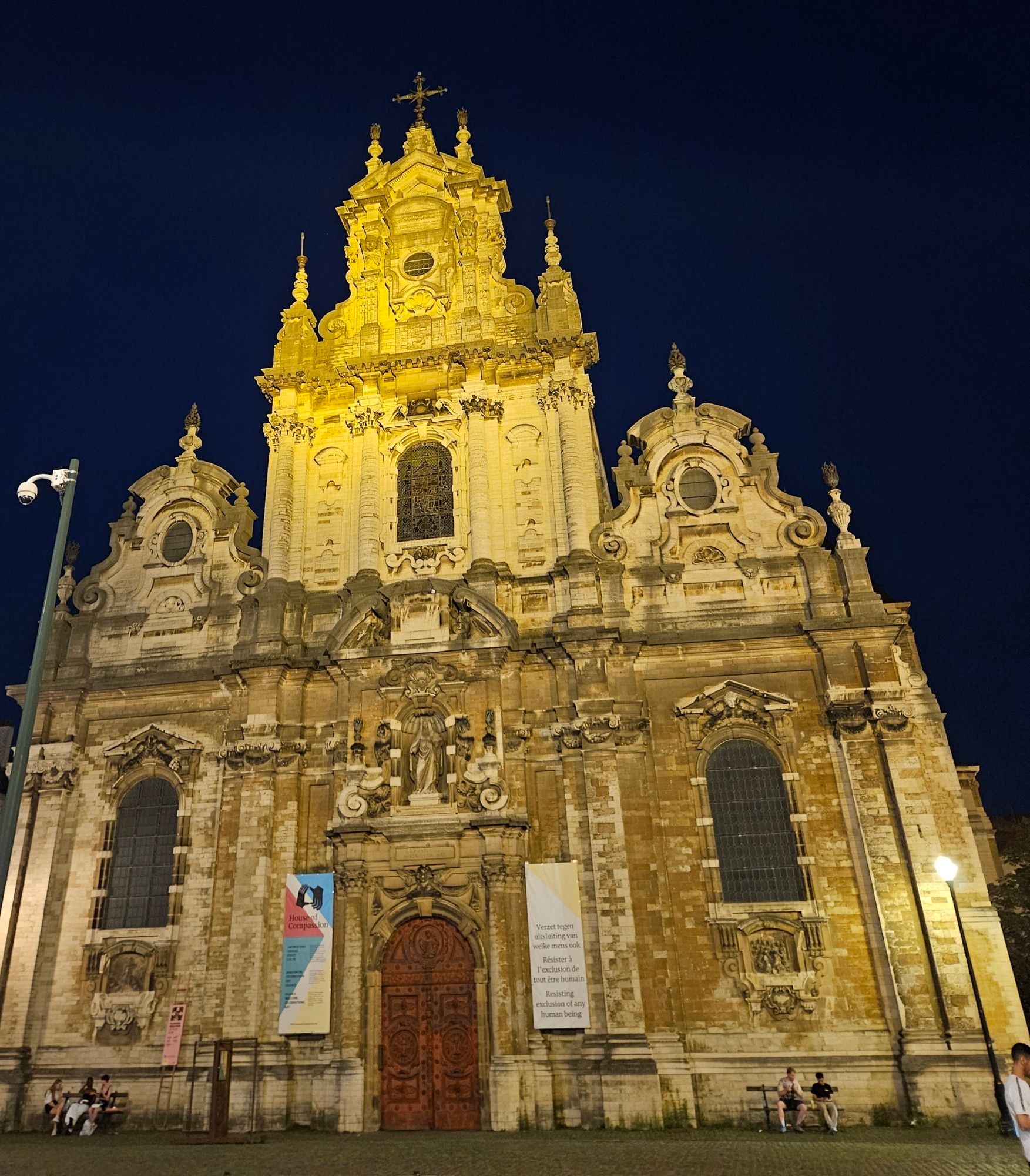 Angestrahlte Barockfassade der Kirche St. Jean Baptise au Béguinage in Brüssel, daran aufgehängt ein Banner mit dem Spruch "Widerstand gegen den Ausschluss jeder Art Mensch" auf Niederländisch, Französisch und Englisch.