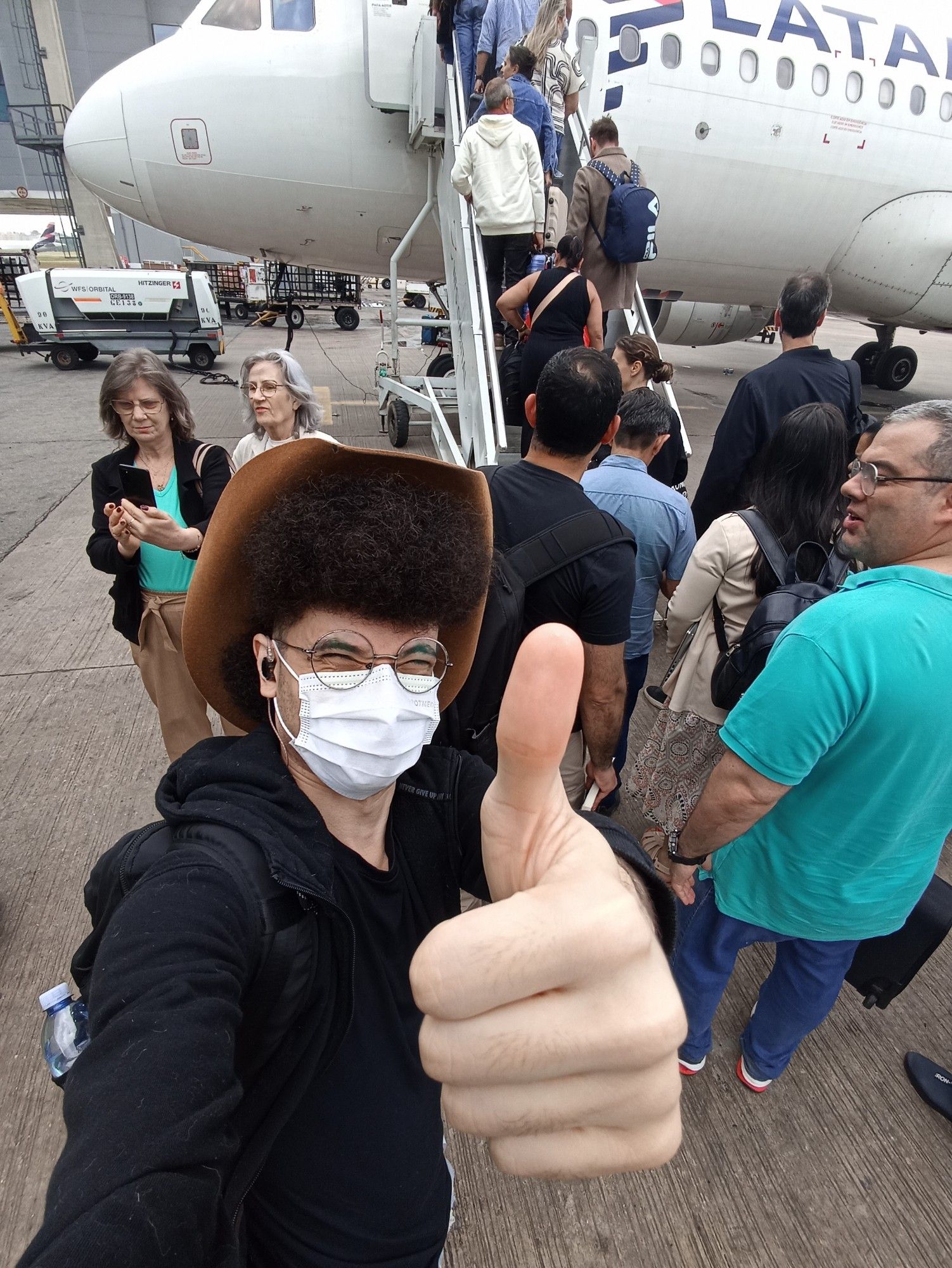 Pessoa com cabelo grande e chapéu de cowboy faz o sinal de jóia para a câmera. No fundo, várias pessoas entrando em um avião.