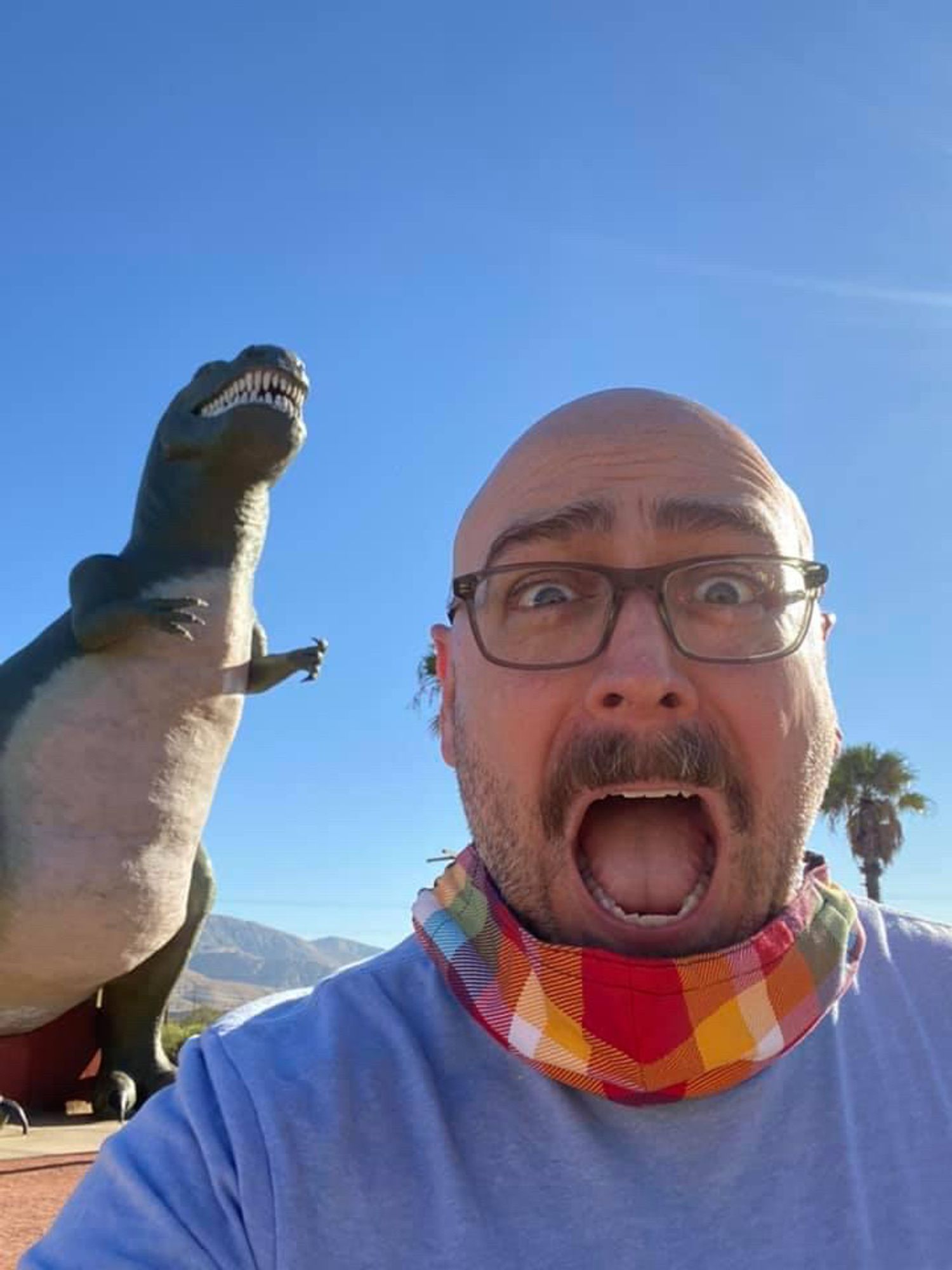 Bald man with mustache and glasses, wearing a light blue shirt with a mask lowered so you can see his mouth open, a look of abject fear in his face. Behind him, a fiberglass T-Rex.