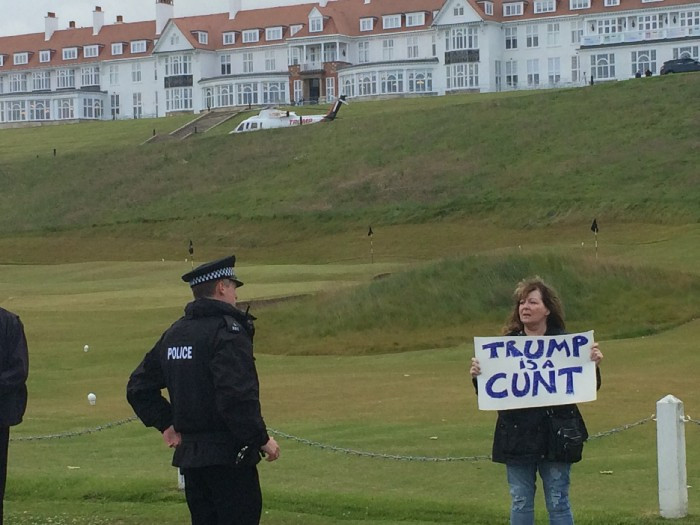 Scottish comedian Janey Godley protests at Trump's golf course in 2016. Standing on the lawn next to a uniformed policeman, she holds a handwritten sign reading Trump is a Cunt.