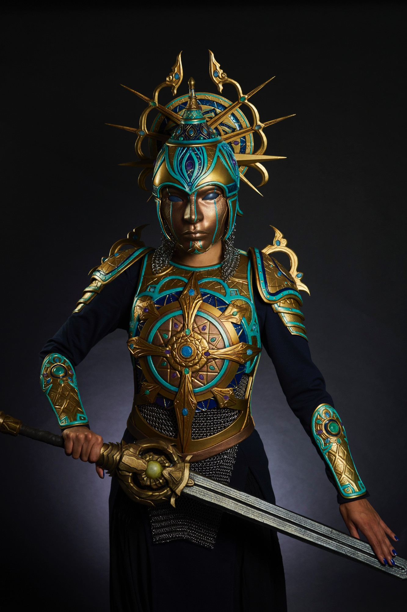 A woman wearing a costume of gold and turquoise armour inspired by traditional Islamic armour. She’s holding a sword.