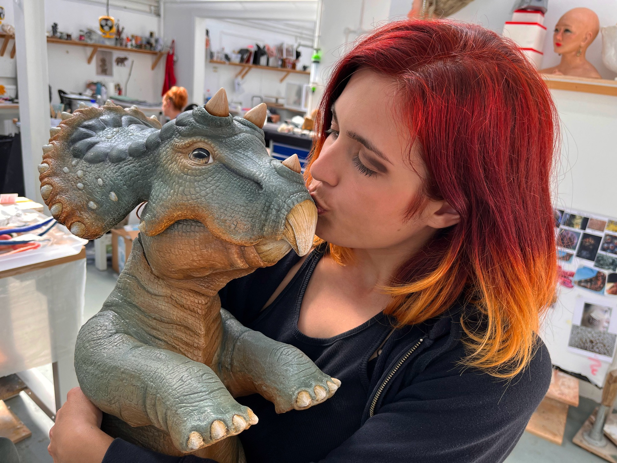 A woman with red and blonde hair holding a baby triceratops puppet with green and tan colouration 