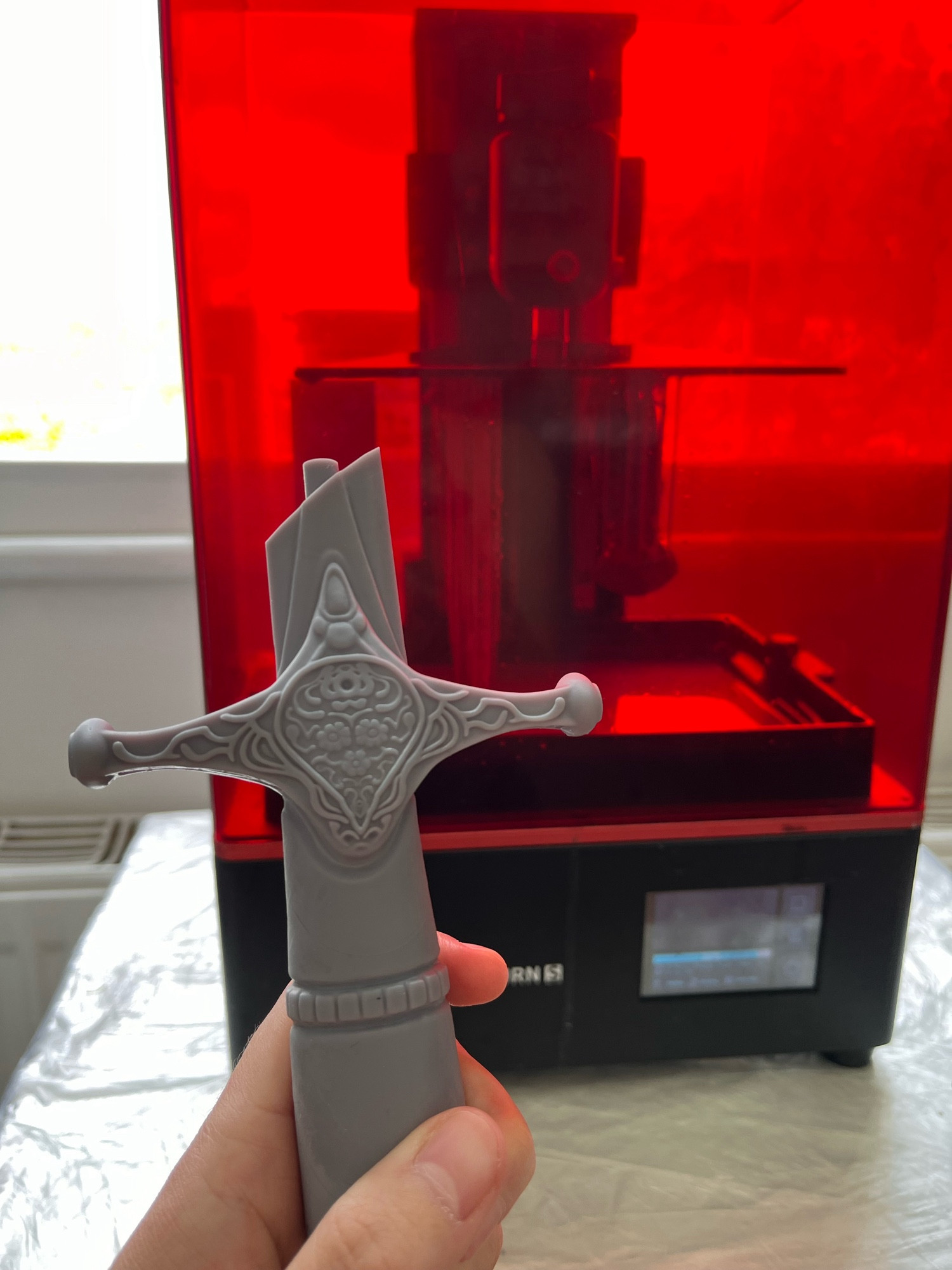 A hand holding up the hilt of a 3D printed dagger. A 3D printer with the other parts in the process of being printed is visible behind it